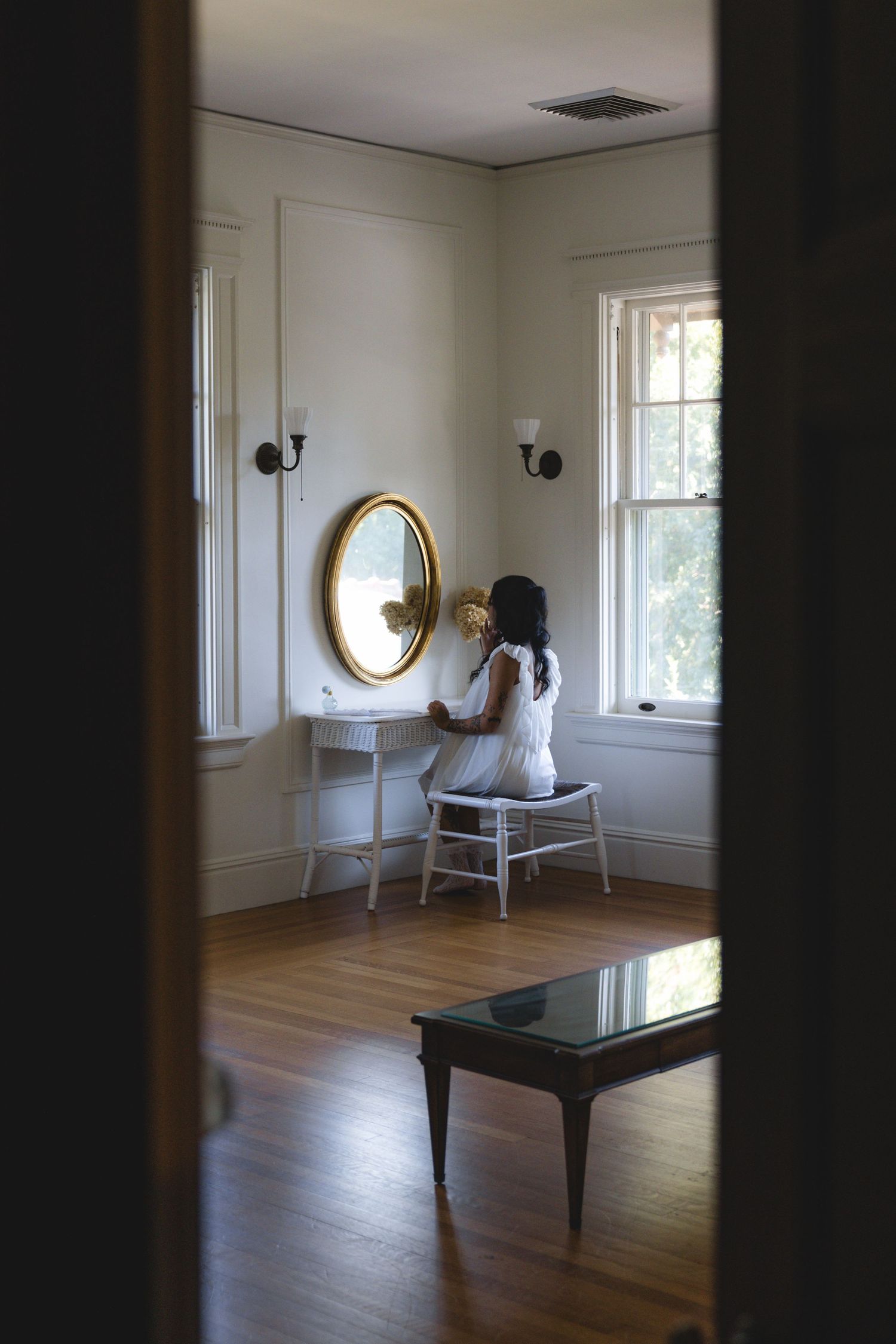 Woman in white dress sitting at vintage vanity mirror in elegant room with hardwood floors and natural light.