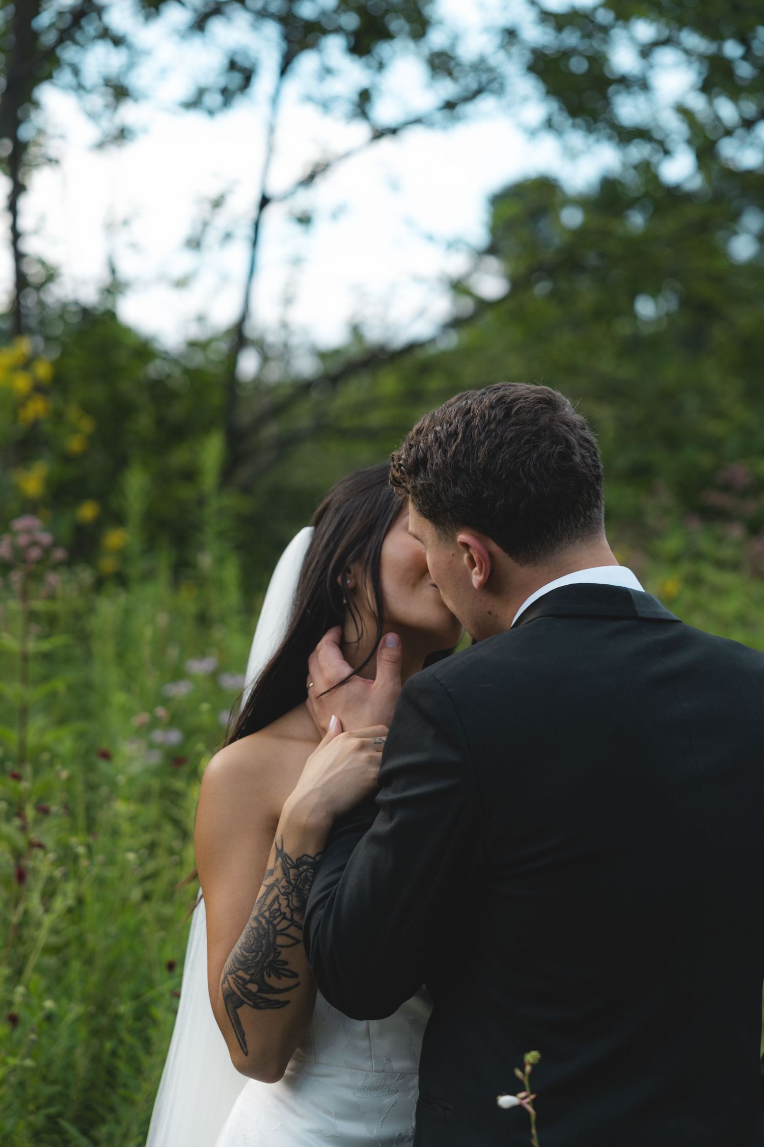 Bride and groom share a romantic kiss outdoors, surrounded by lush greenery, groom's tattooed hand cradling her face.