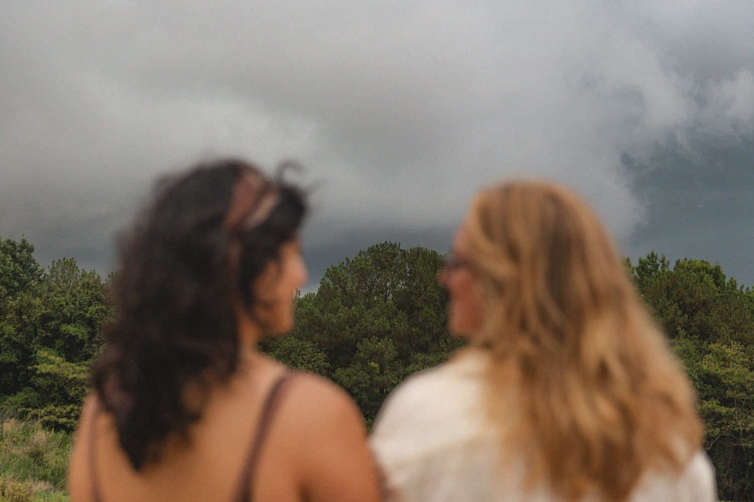 Two women with curly and blonde hair stand outdoors under dramatic storm clouds with lush green trees behind them.