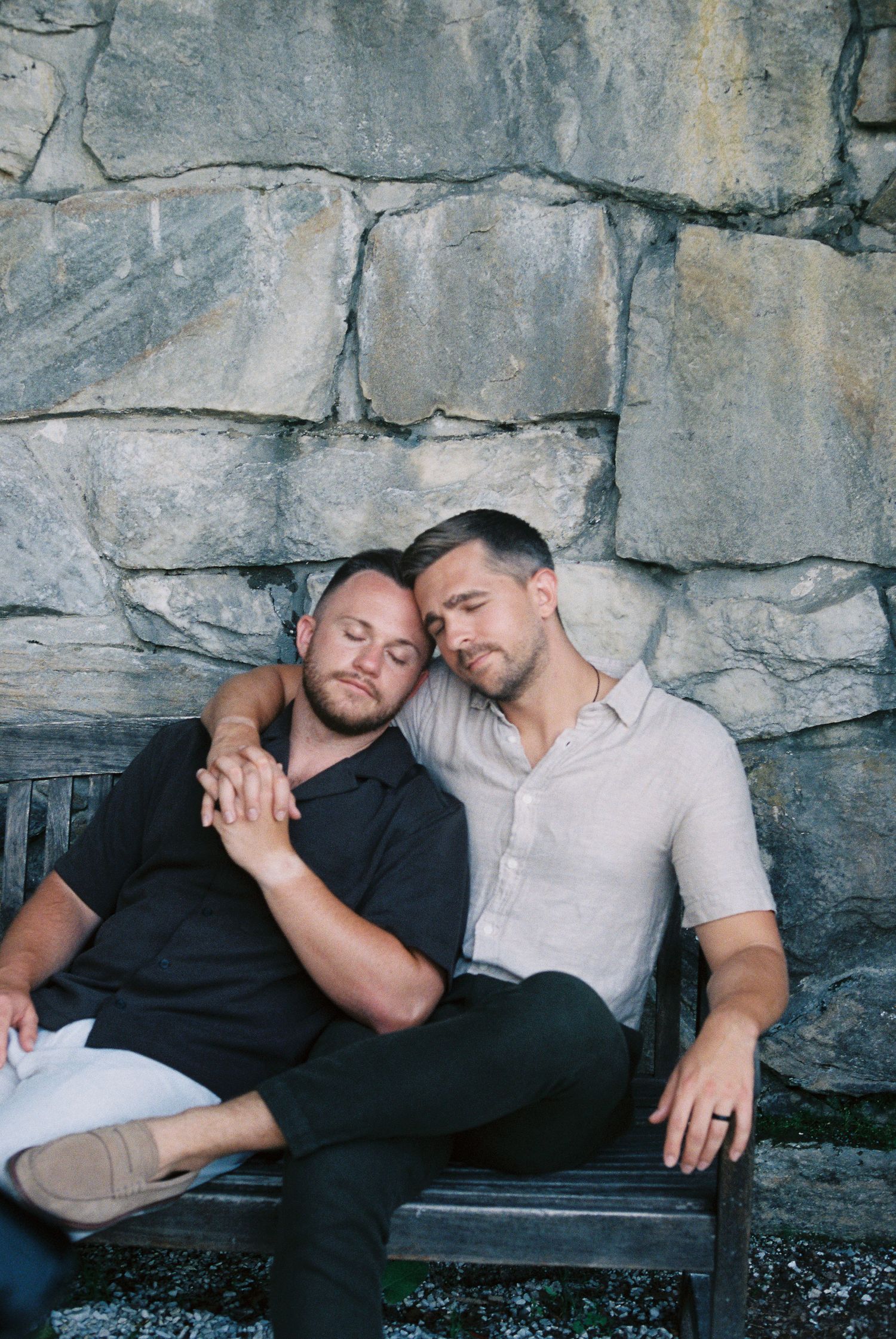 Two men sitting on a bench, leaning on each other affectionately against a stone wall backdrop.