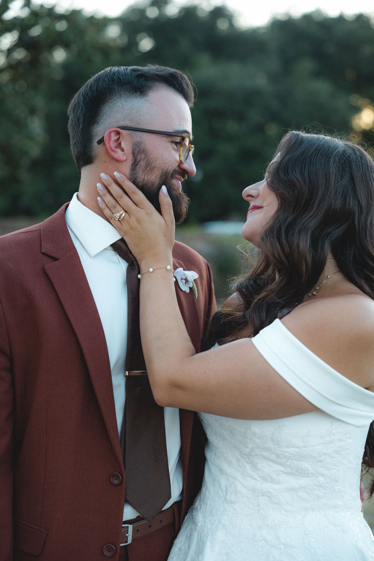 A bride in a white off-shoulder gown tenderly touches the face of her groom in a brown suit during their wedding.
