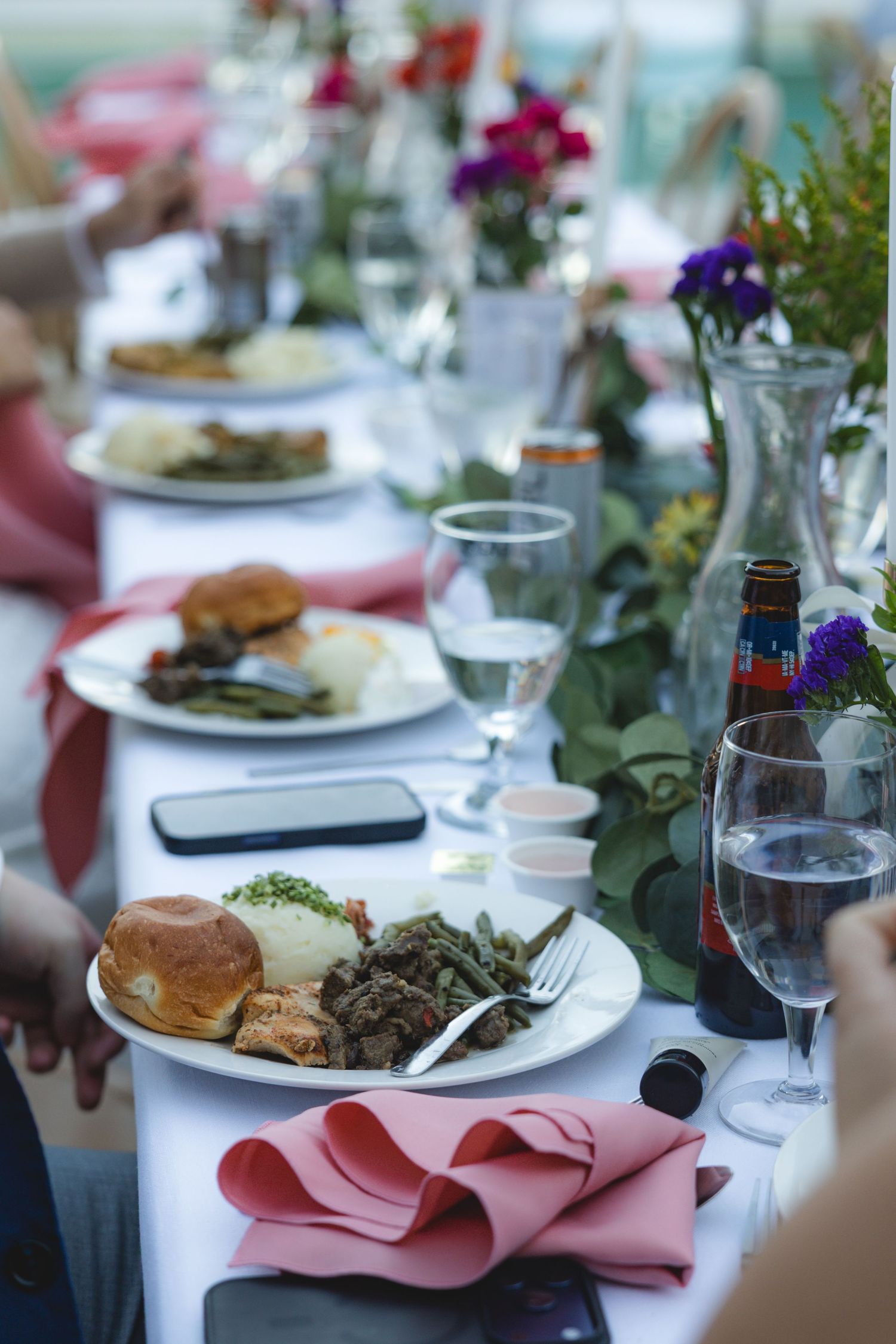 Elegant wedding reception dinner table with plates of food, pink napkins, floral centerpieces, and glassware.