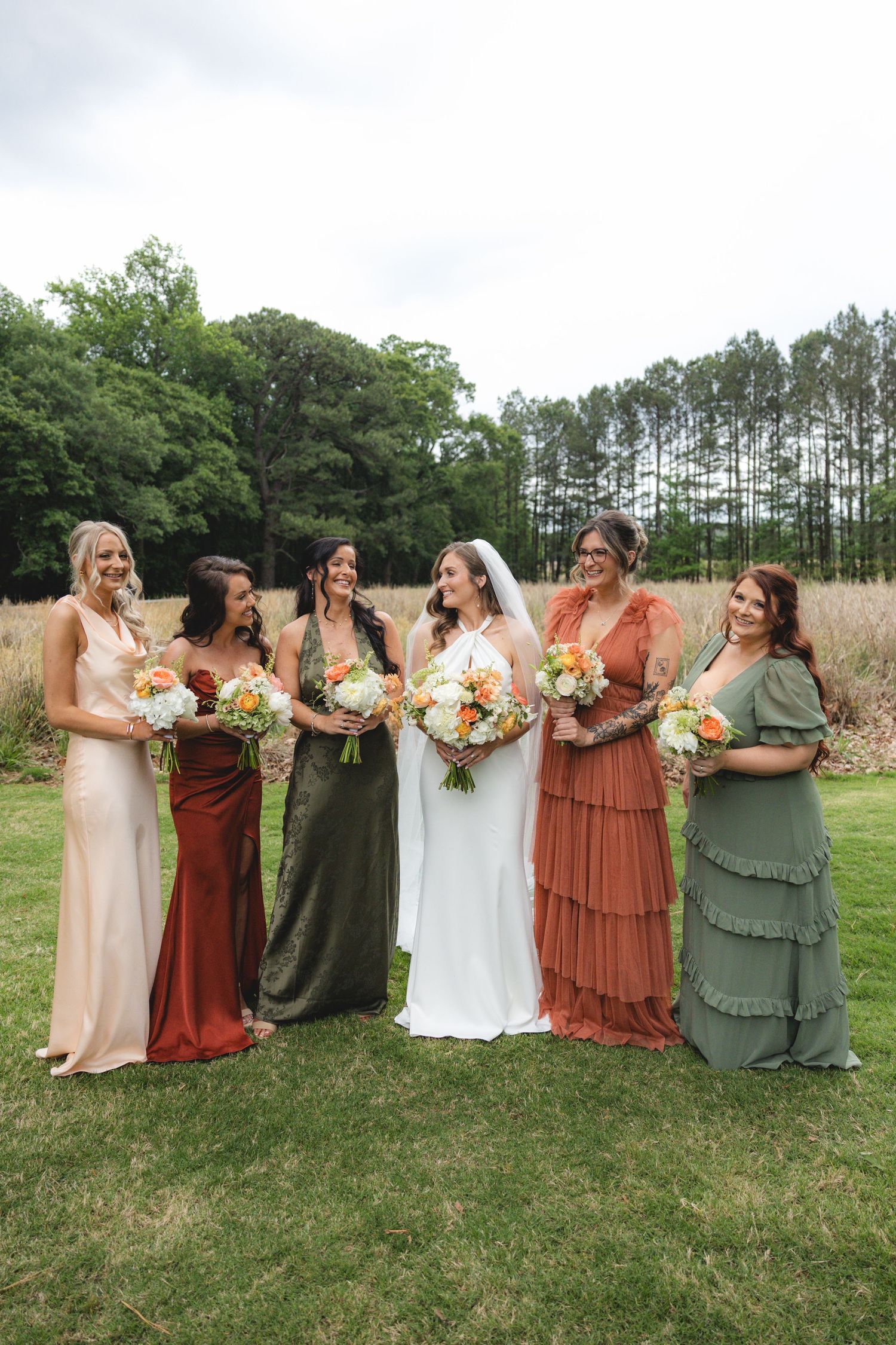 Bride with bridesmaids in mismatched earth-tone gowns holding colorful bouquets laughing outdoors.