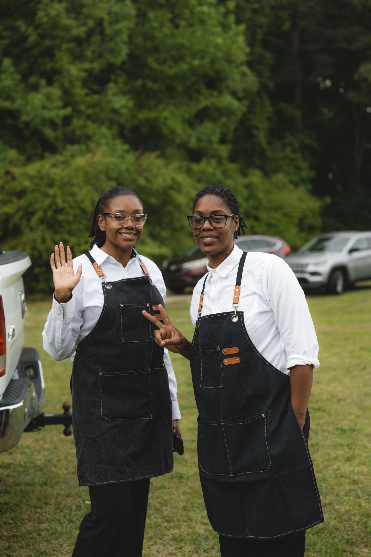 Two female catering staff in black aprons smiling and waving outdoors.