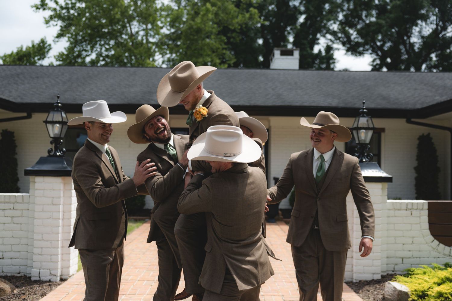 Groomsmen in brown suits and cowboy hats celebrate with joyful embrace outside white building.