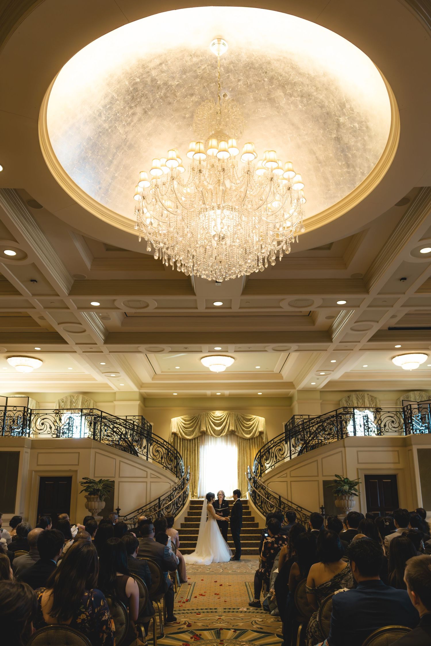 Wide angle view of wedding ceremony under crystal chandelier with grand dual staircase backdrop.