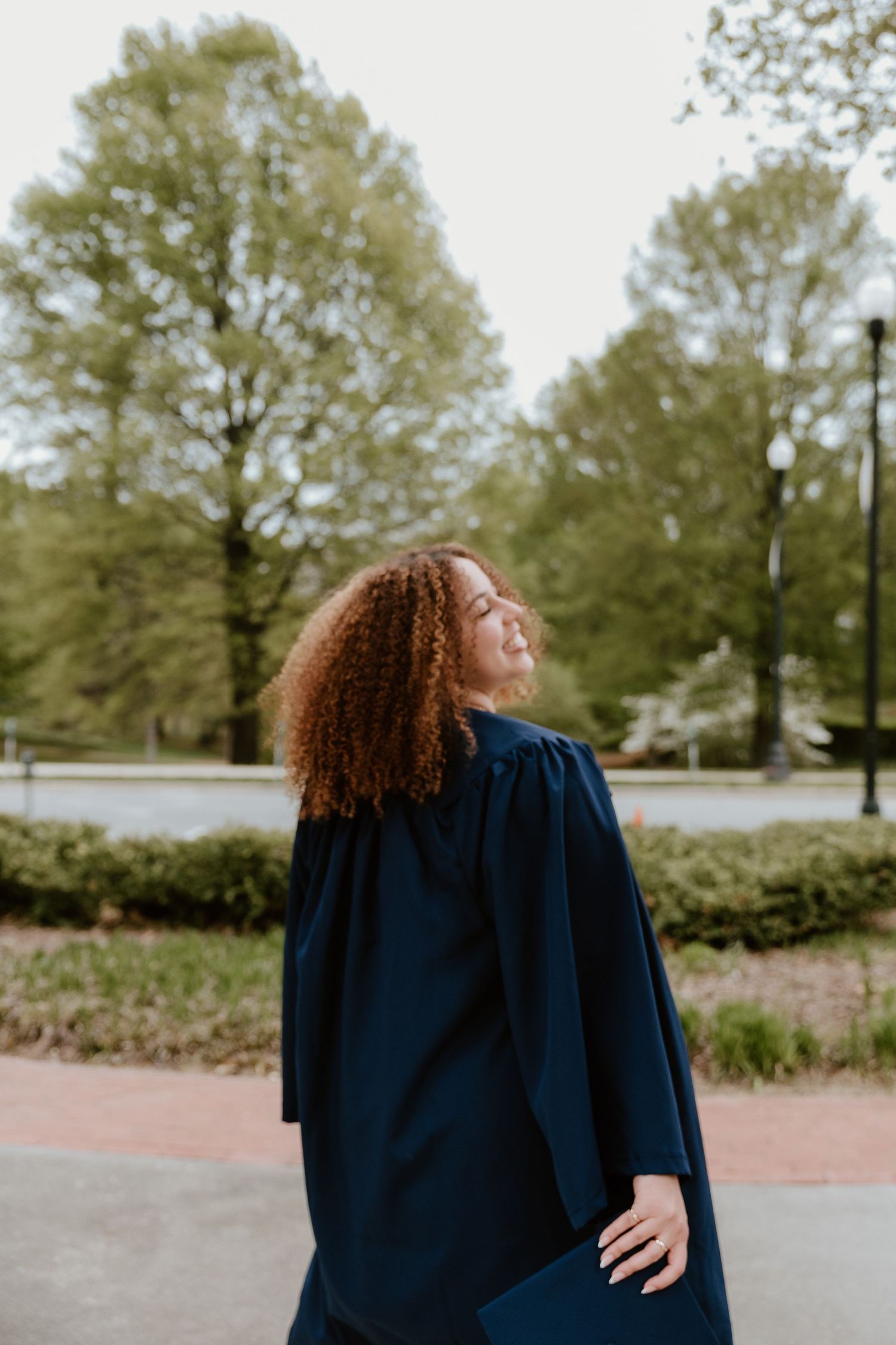 Young woman with curly red hair wearing a navy graduation gown poses outdoors on campus.
