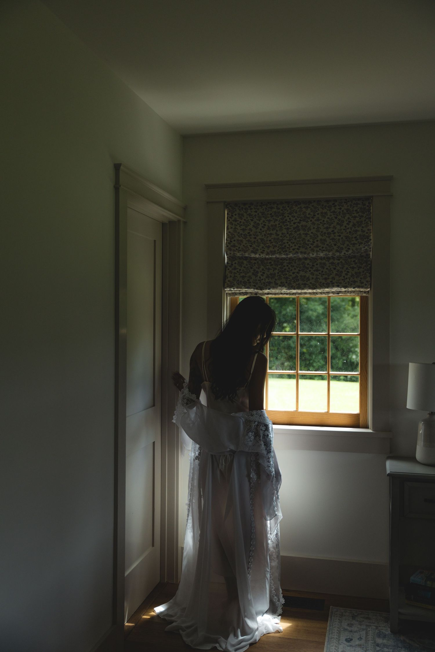 A bride in a white wedding dress stands silhouetted by a sunlit window in a softly lit room.