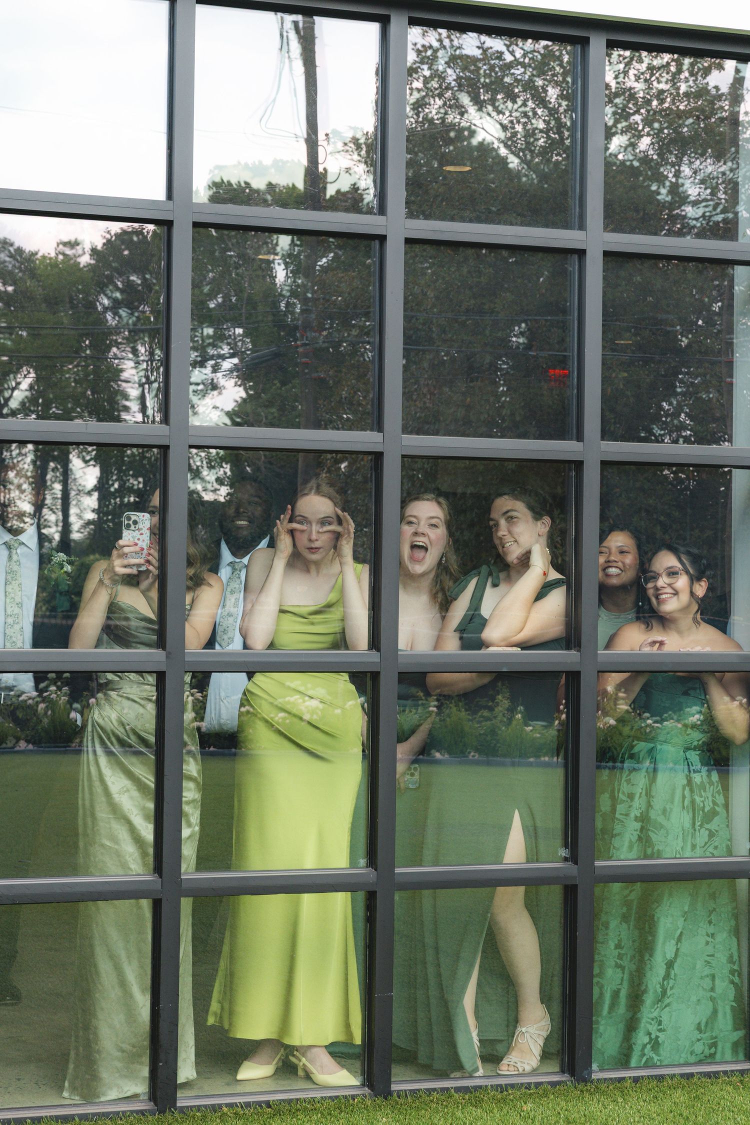 A group of young women in elegant green formal gowns posing and laughing behind a large grid window.