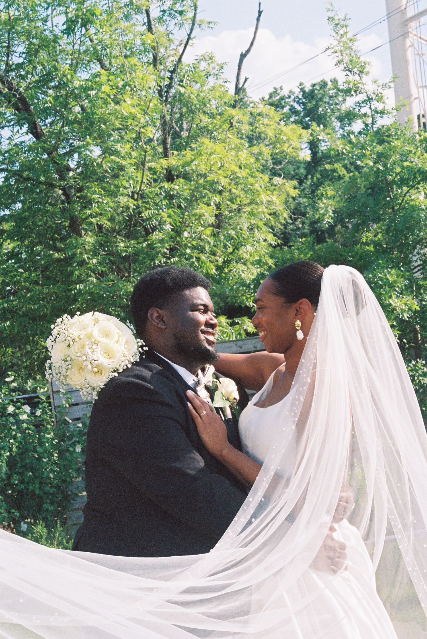 A joyful bride and groom share a romantic moment outdoors, her white veil flowing, surrounded by lush green trees.
