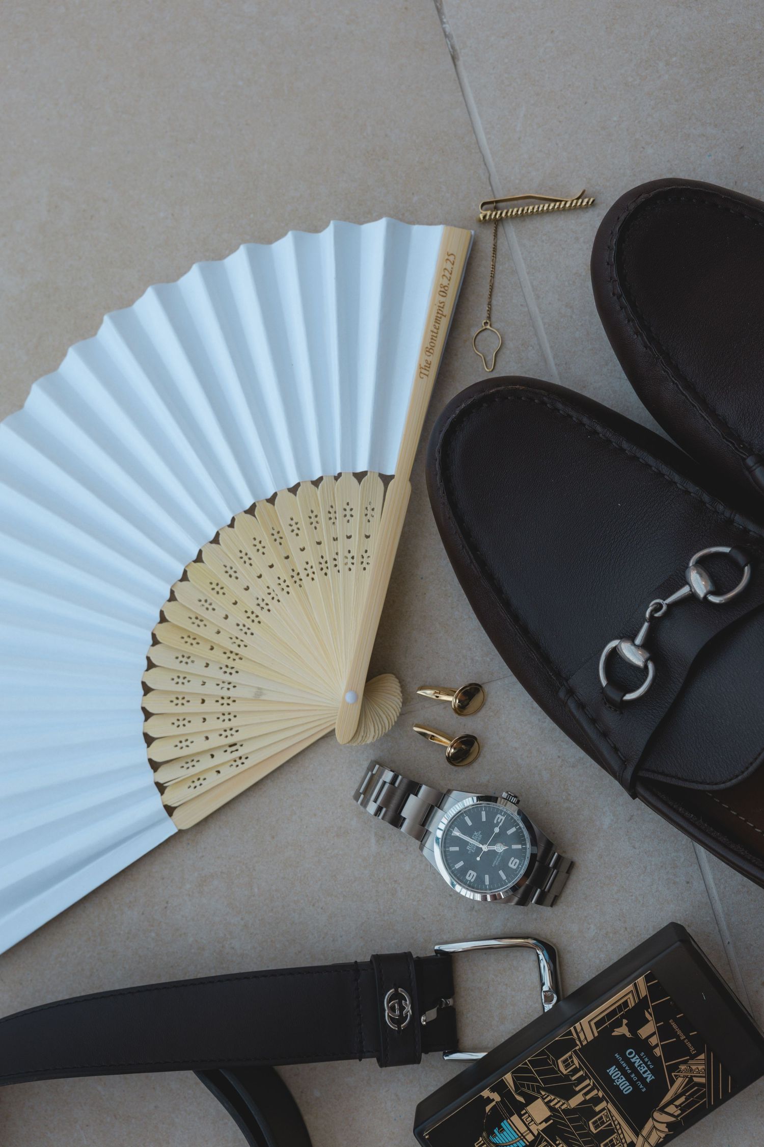 Flat lay of men's accessories: white hand fan, loafers, watch, cufflinks, leather belt, and wallet on gray surface.