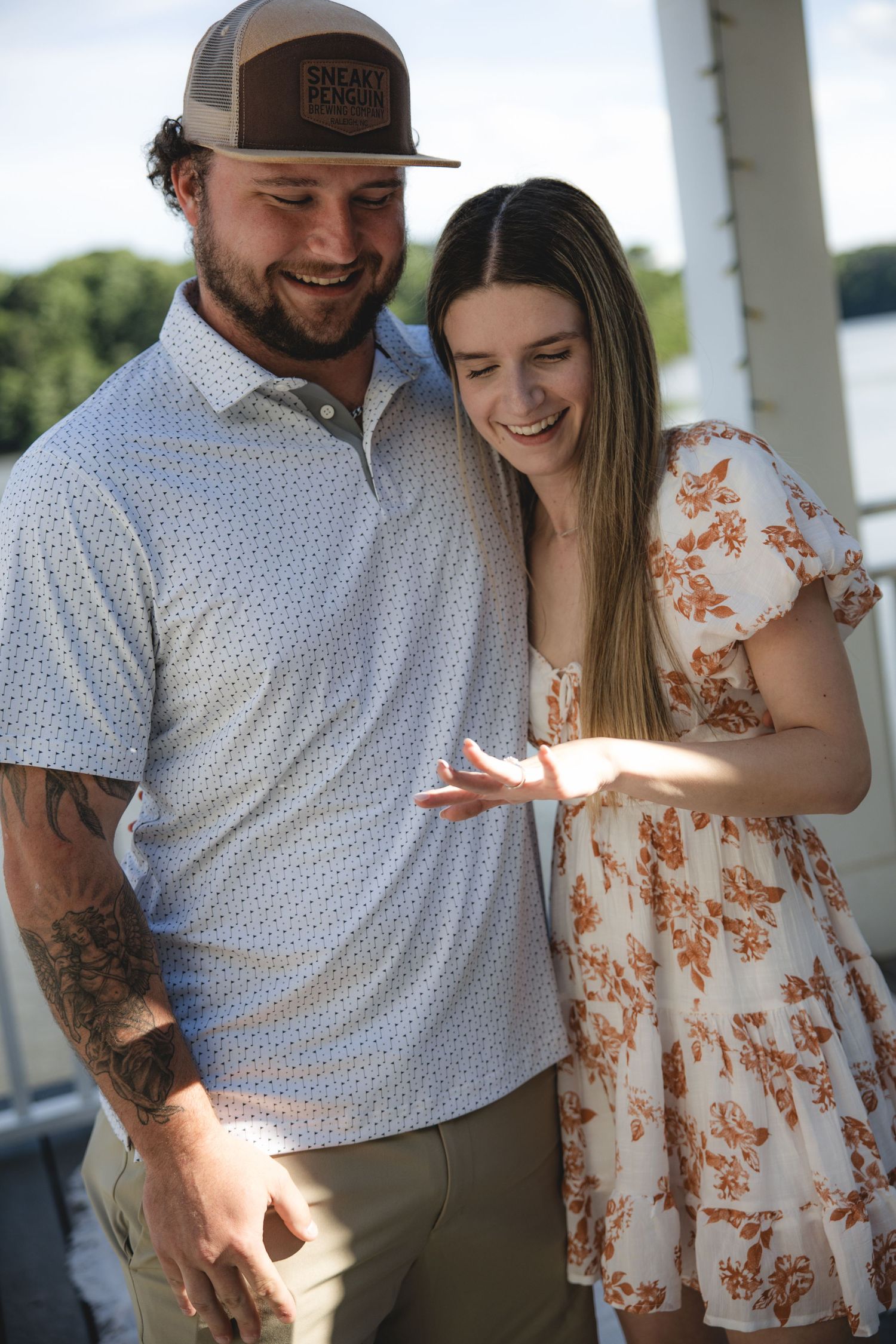Happy couple smiling and admiring an engagement ring outdoors near a lake, man with tattoos in polo shirt and cap.