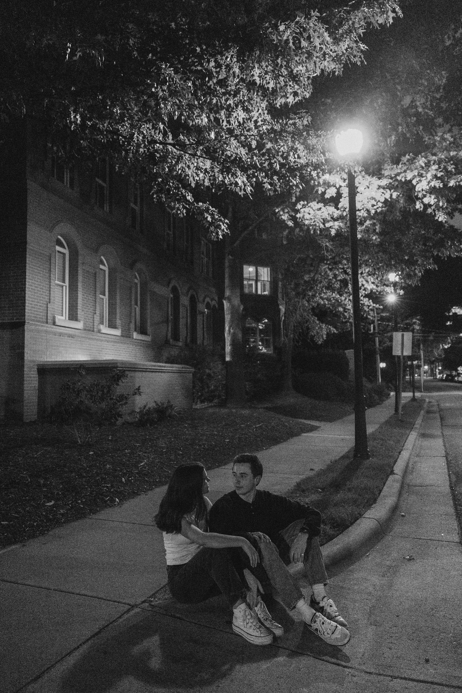 Black and white photo of a couple sitting on a curb at night under a street lamp beside a brick building.
