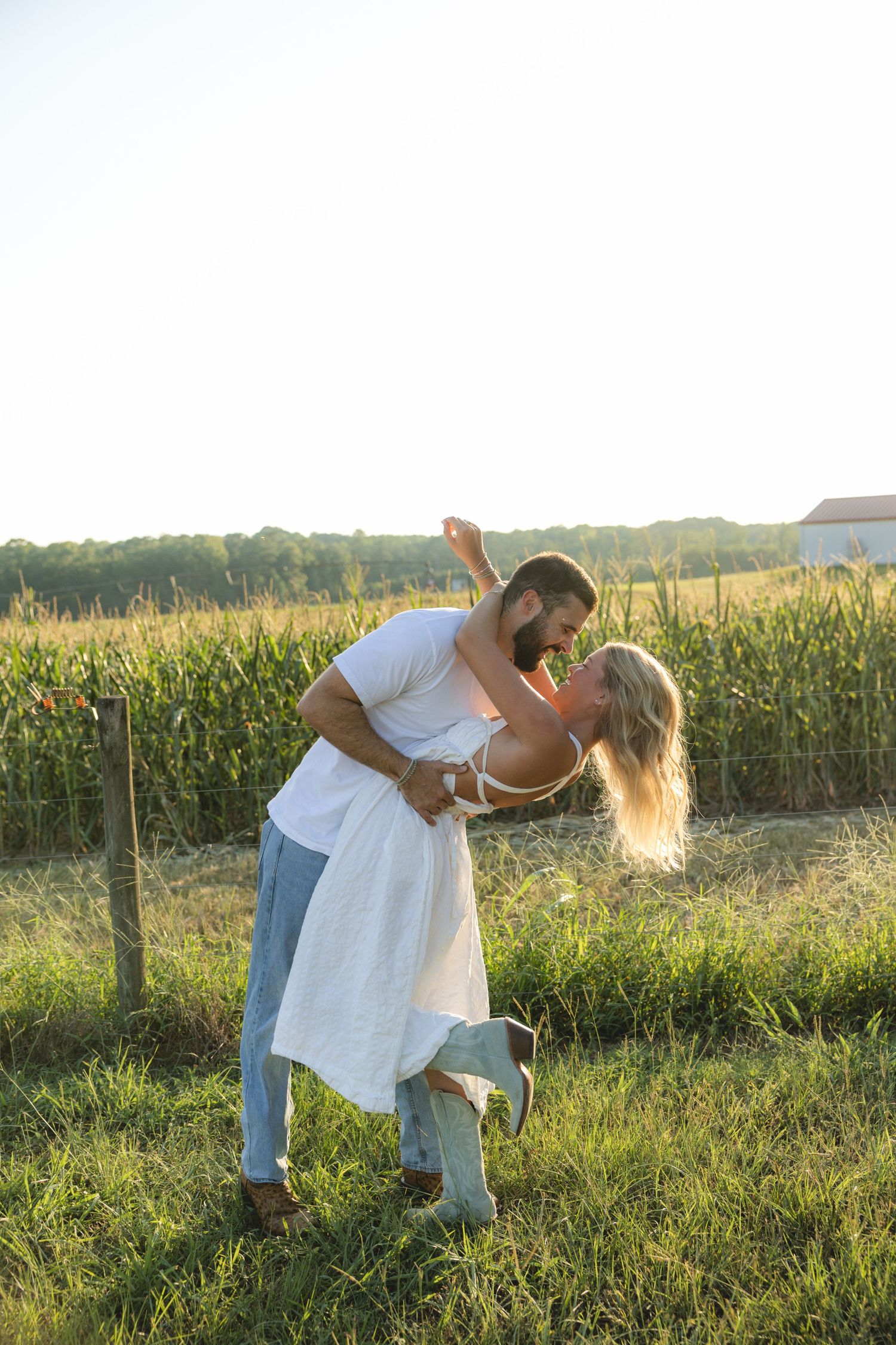 Couple dancing and almost kissing in a sunny green field near a cornfield, man dipping woman in white dress.