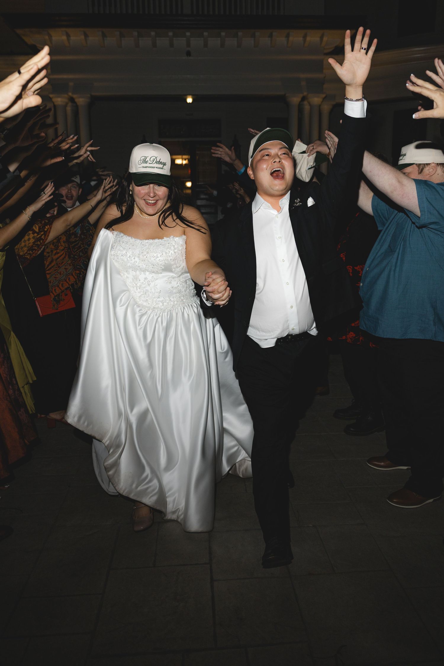 Bride in a white satin gown and groom in a black suit, both wearing caps, hold hands and cheer as guests raise their arms at a wedding.