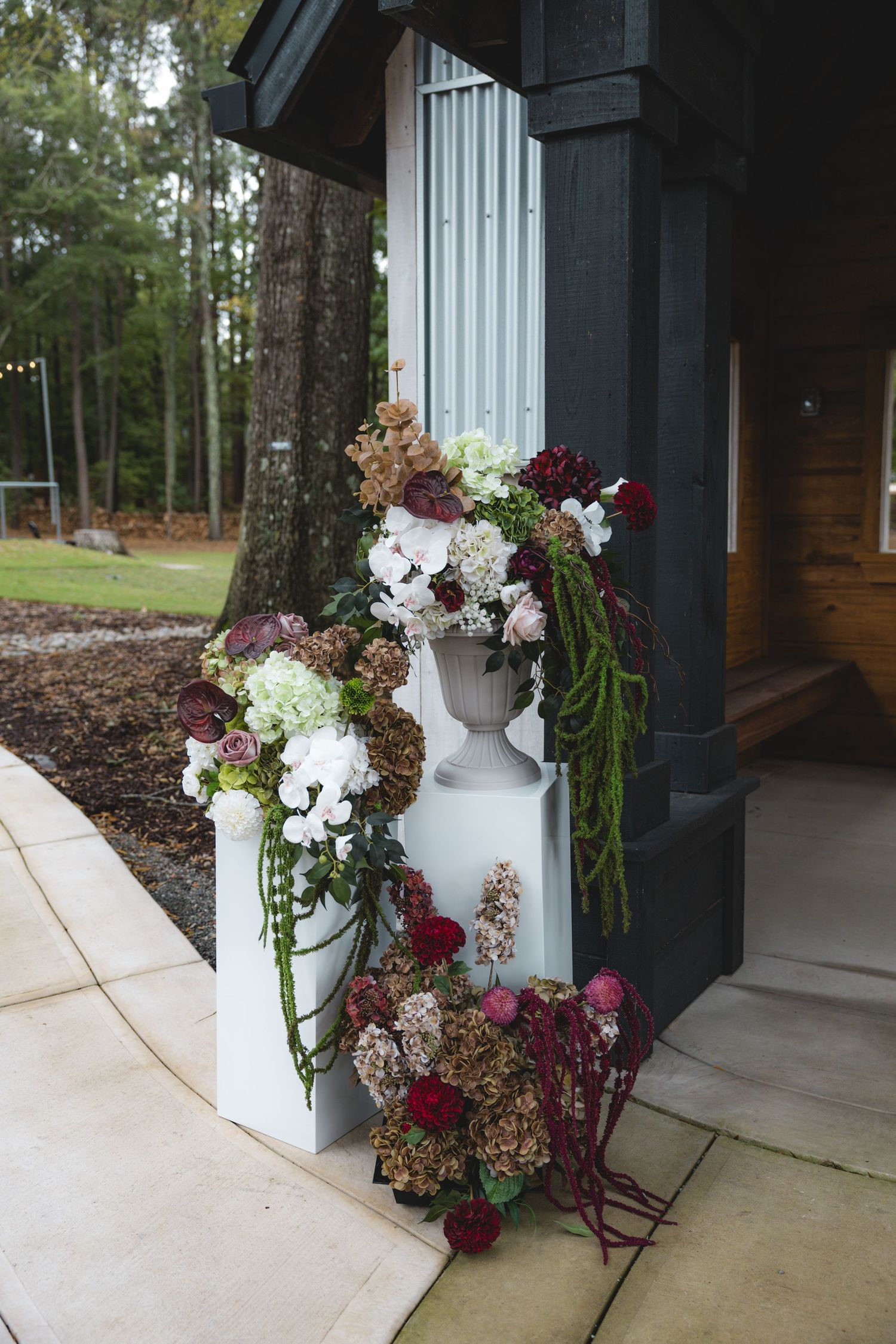 Burgundy, blush and white floral arrangements with trailing greenery on white pedestals beside a dark wood porch at a rustic wedding venue.