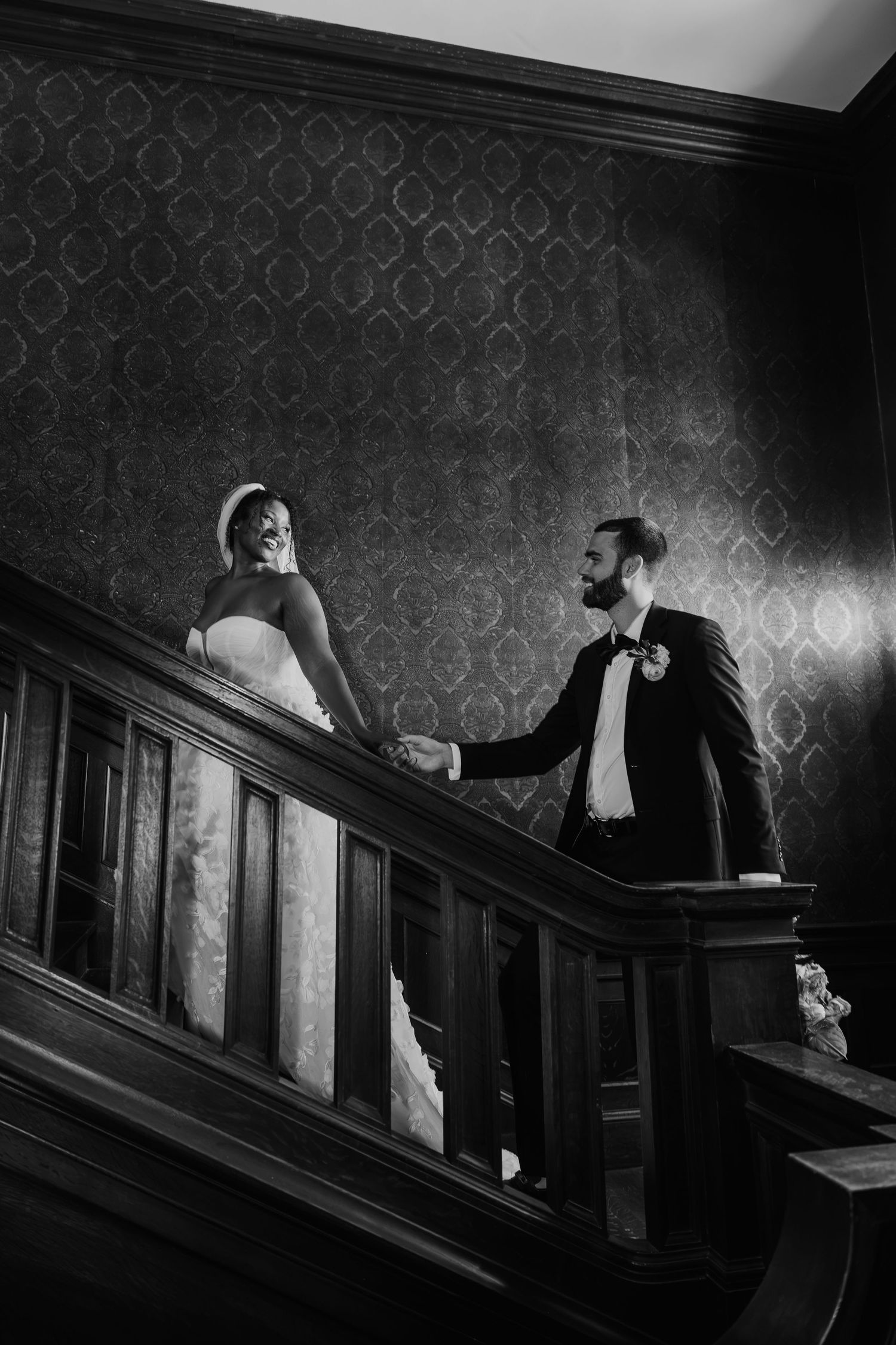 Black and white photo of bride and groom holding hands on elegant wooden staircase with ornate wallpaper backdrop.