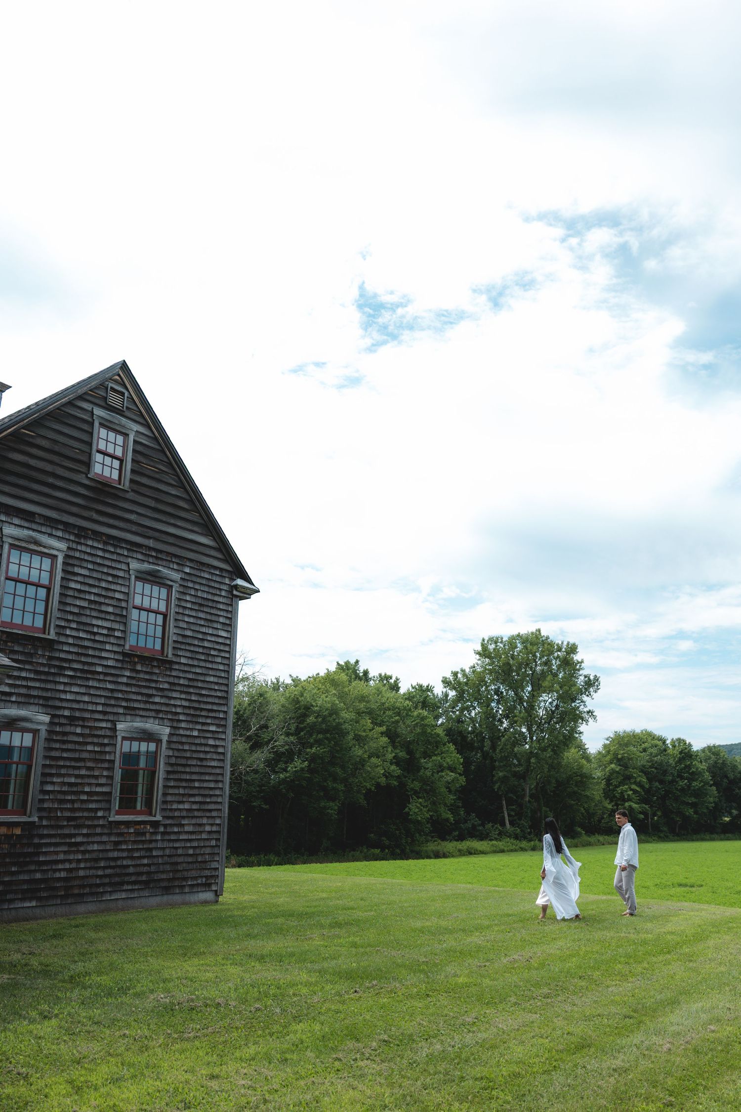 A bride and groom walk hand in hand across a green lawn beside a rustic wooden building with trees in the background.