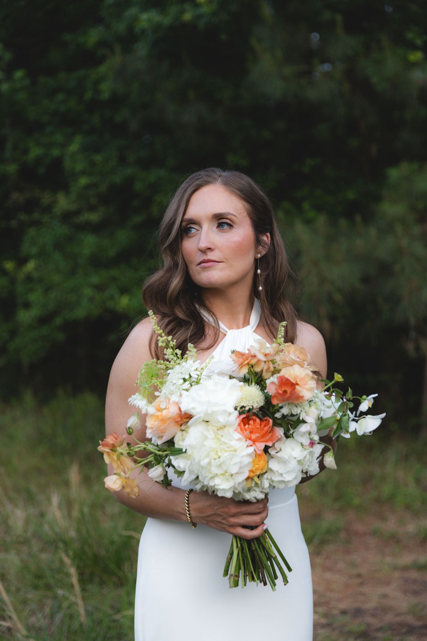 Bride in white halter gown holds white and orange bouquet of peonies and roses, gazing upward outdoors.