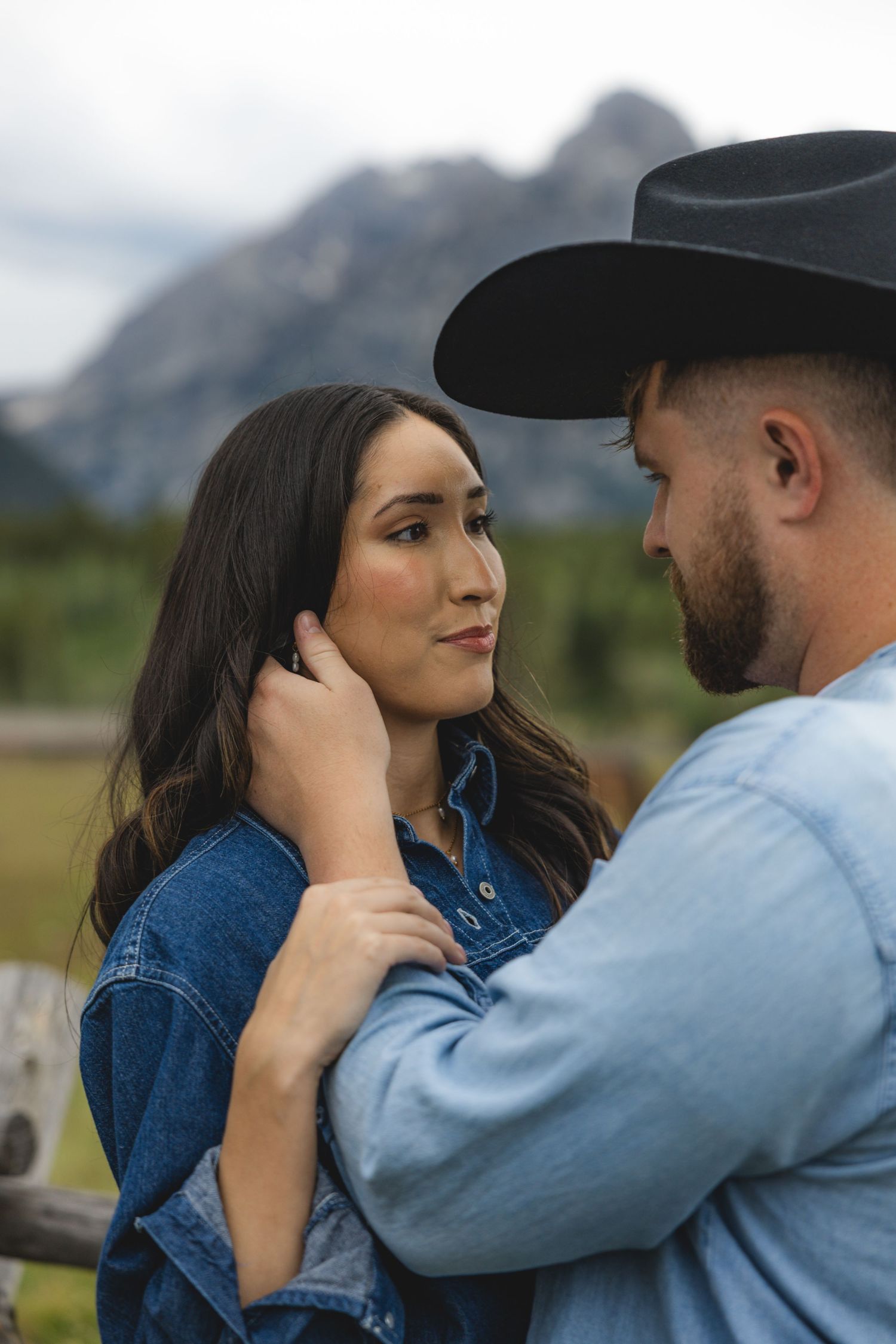 A couple in denim shares a tender moment with mountain scenery, man in black cowboy hat gently touching woman's face.