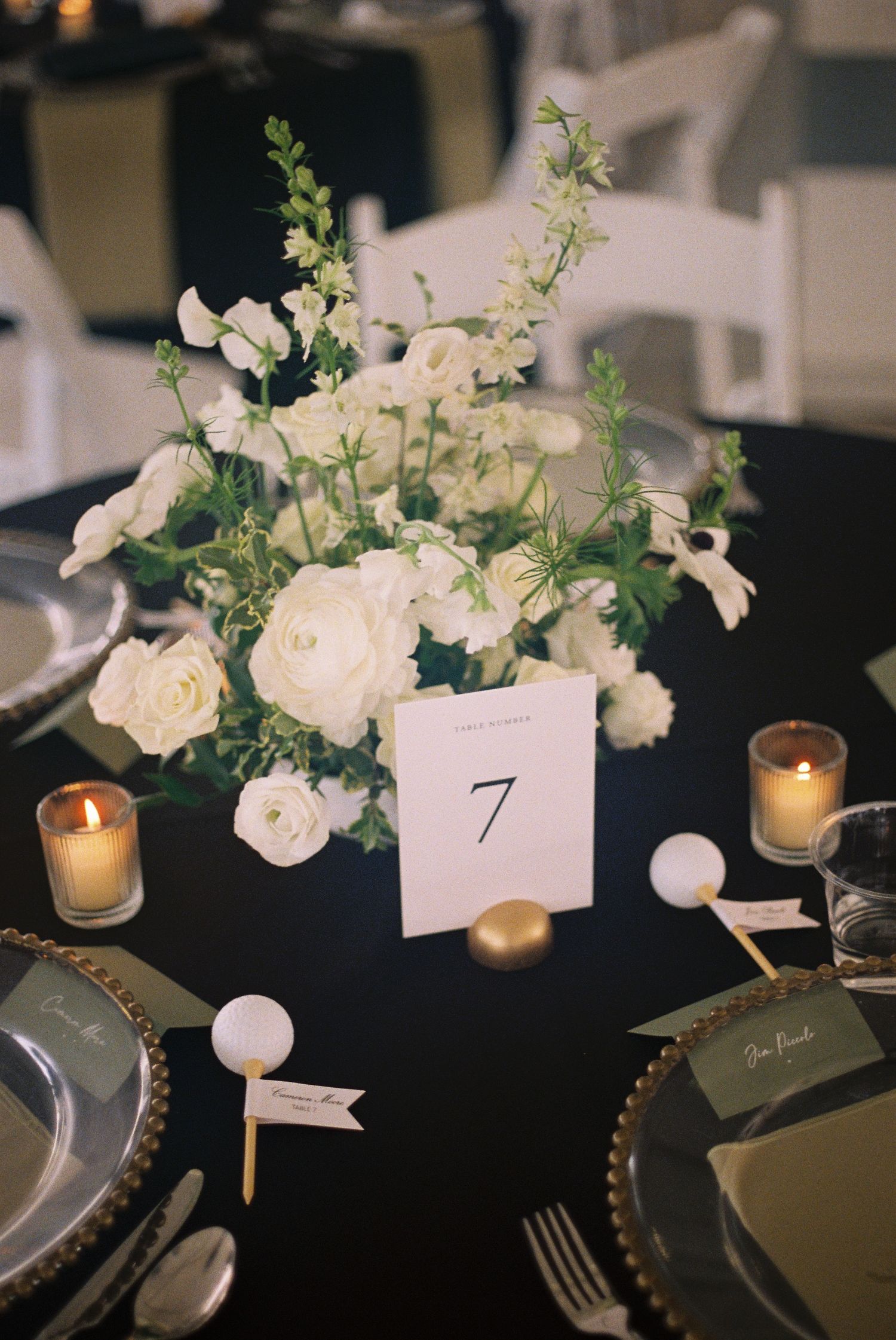 Elegant wedding table setting with white floral centerpiece, gold candles, and table number 7 on black linen.