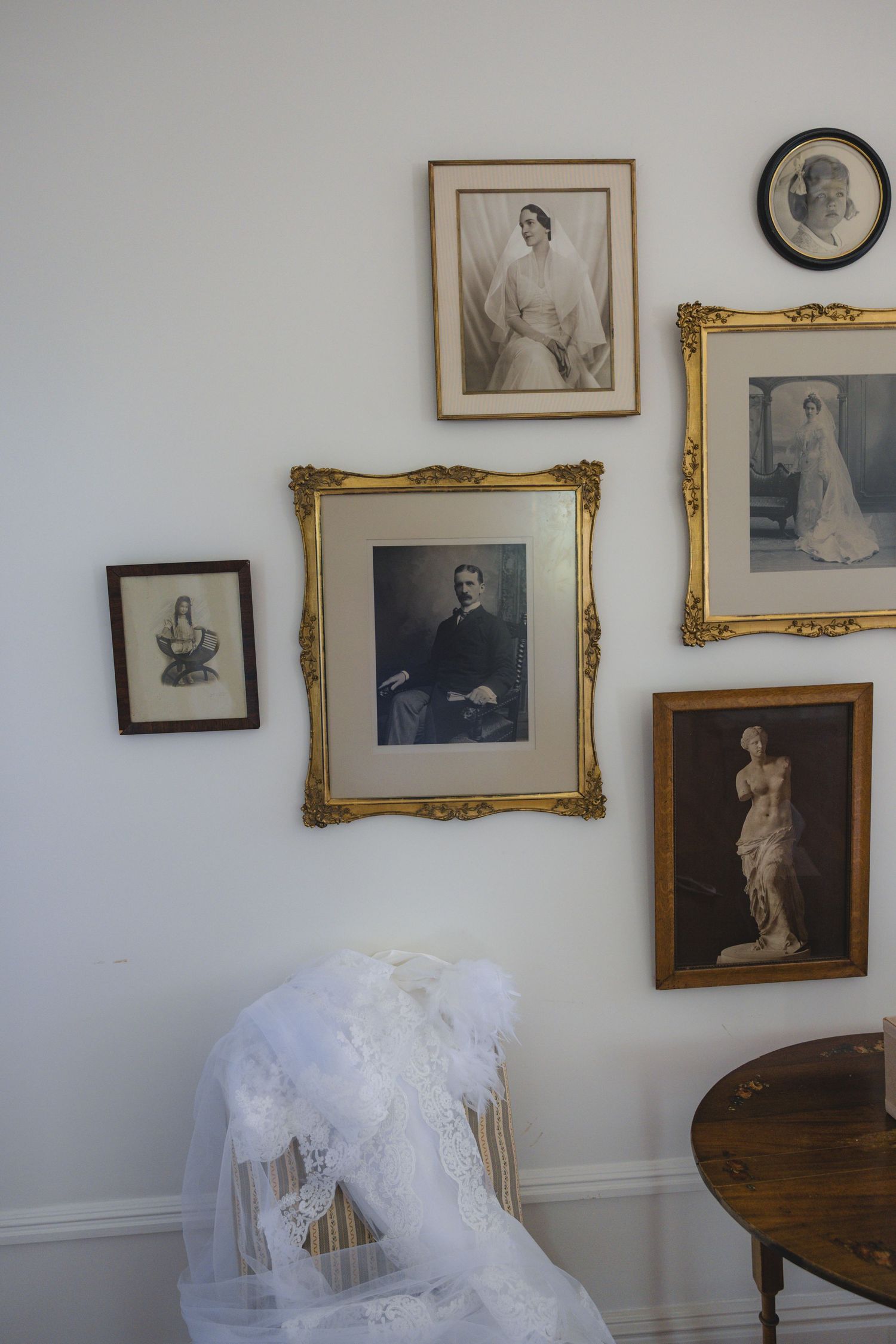 Vintage gallery wall with antique framed portraits and a sculpture photo above a white veiled chair and wooden side table.