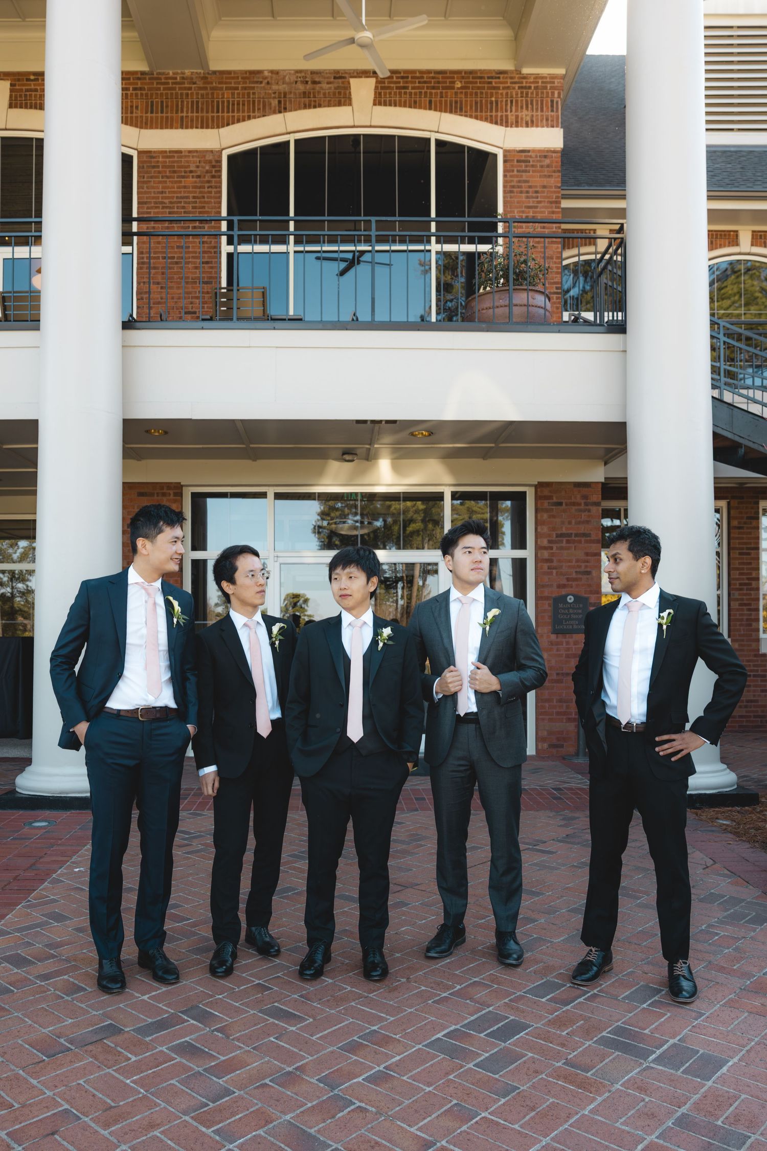 Five groomsmen in black suits with white ties posing together outside brick wedding venue columns.