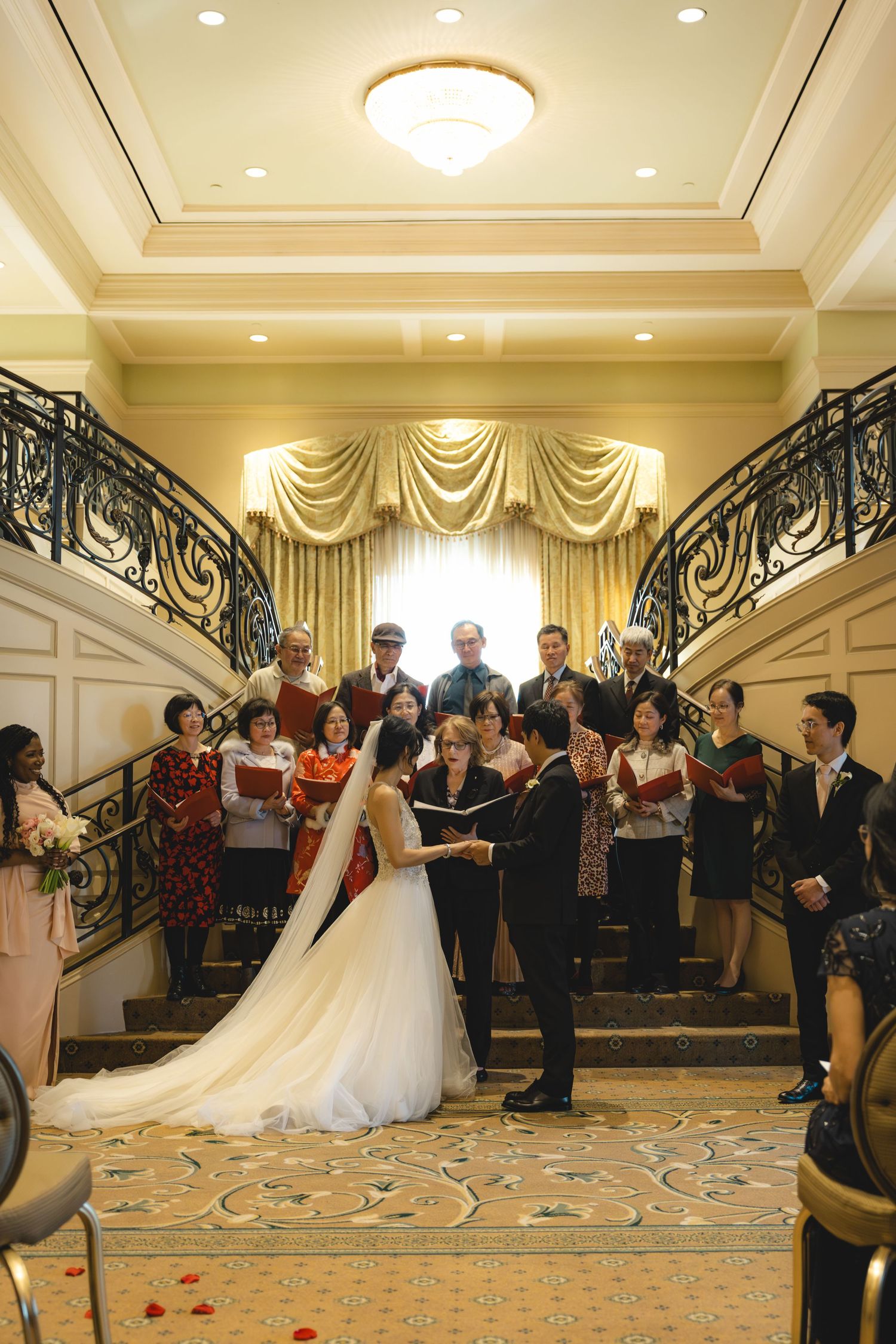 Couple exchanges vows before choir on grand staircase in elegant ballroom wedding ceremony.