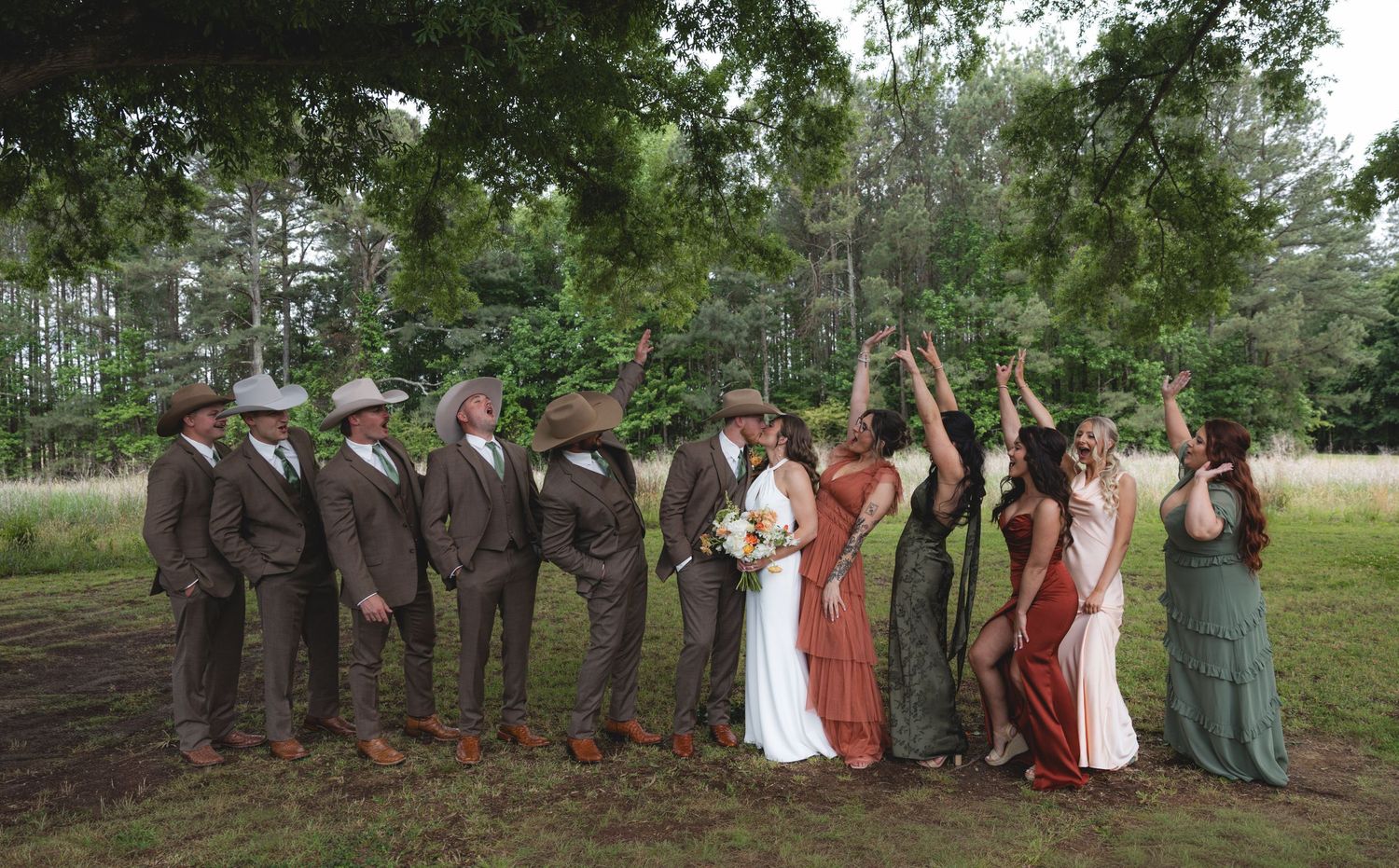 Wedding party poses under large oak tree with groomsmen in brown suits and bridesmaids in earth-tone dresses.