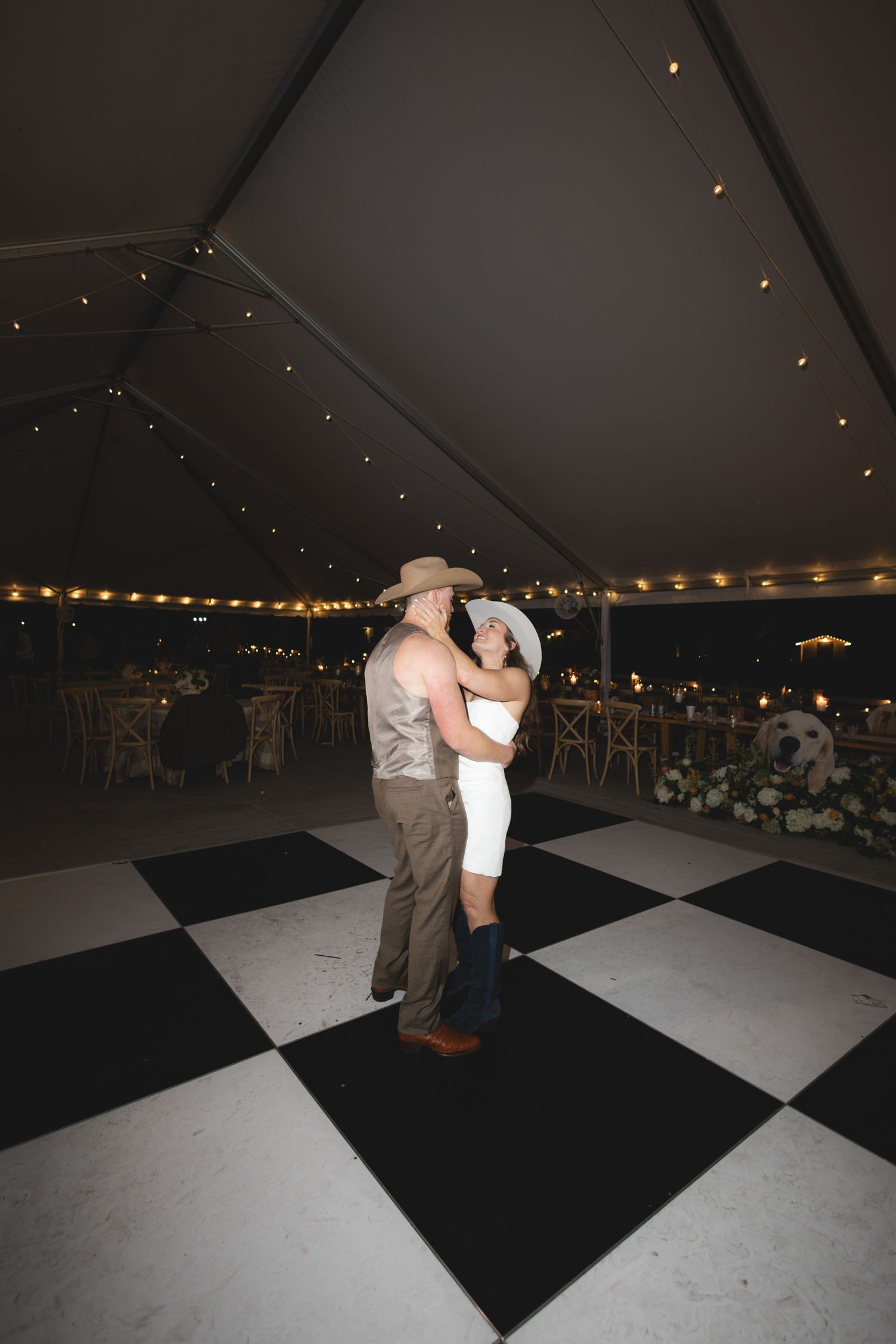 Bride and groom in cowboy hats sharing a slow dance on a checkered floor inside an elegant wedding tent.