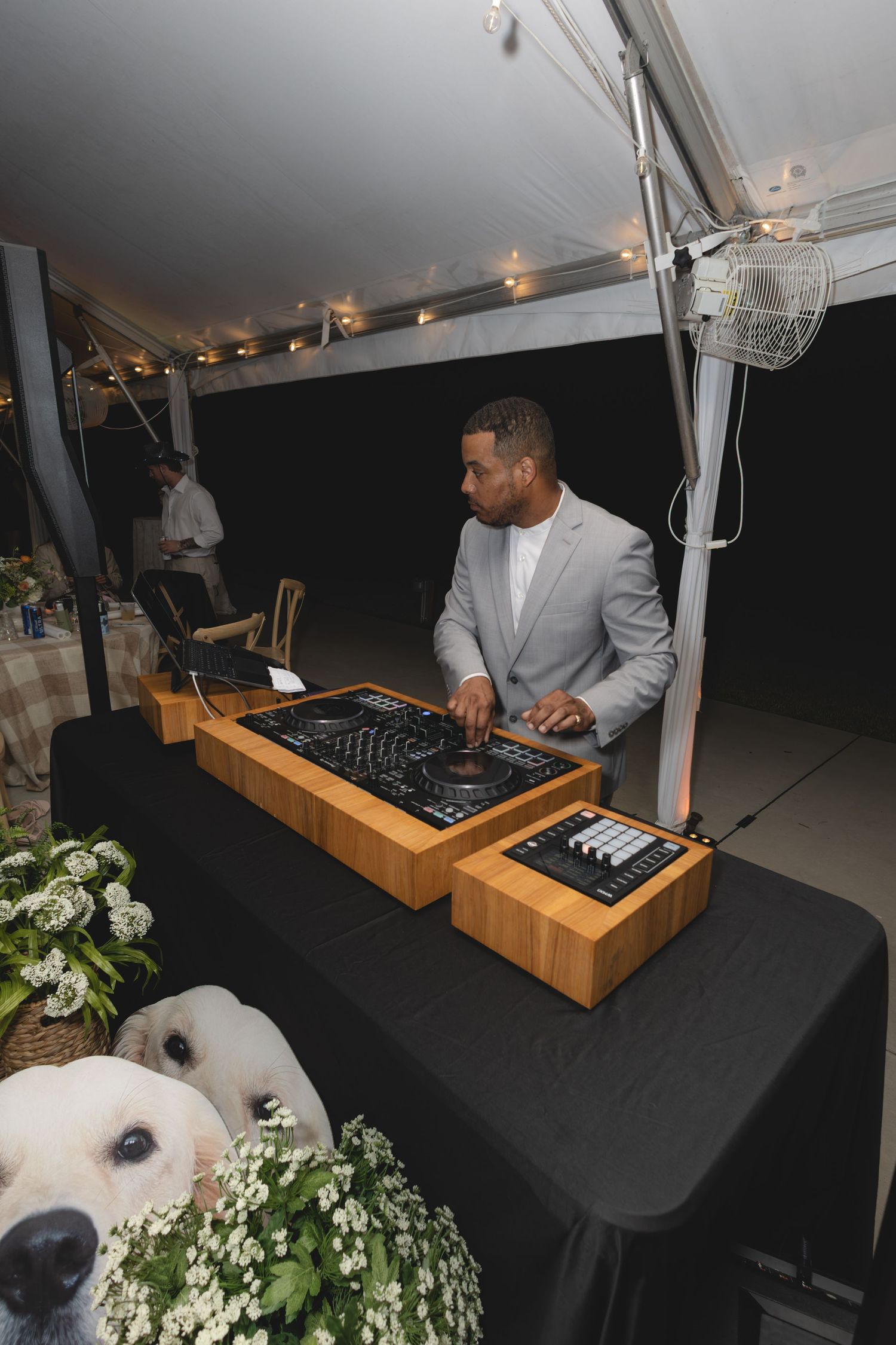 DJ in gray suit mixing music at a wedding reception under a tent.