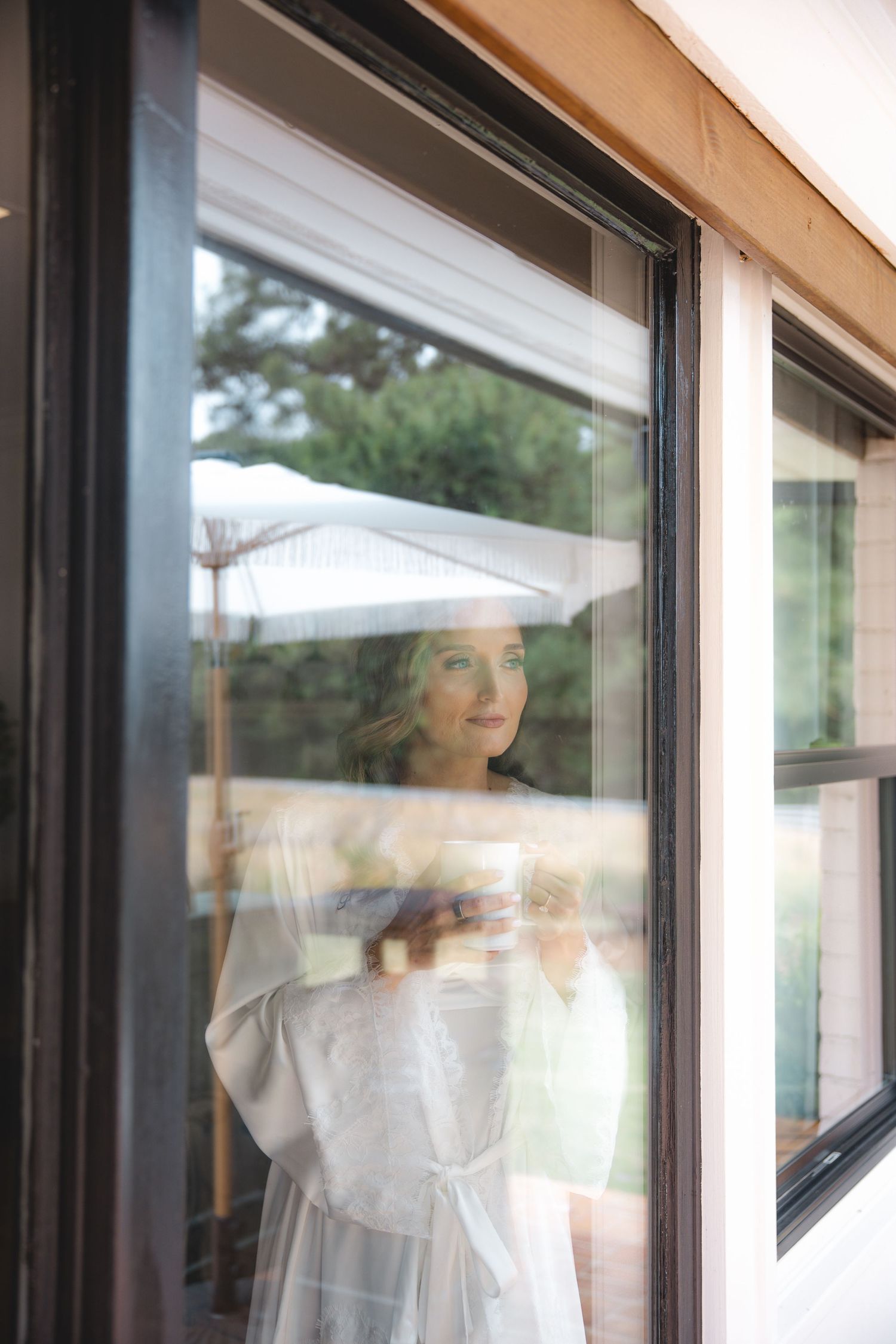 Bride in white robe smiles while looking through large window reflecting outdoor greenery and umbrella.