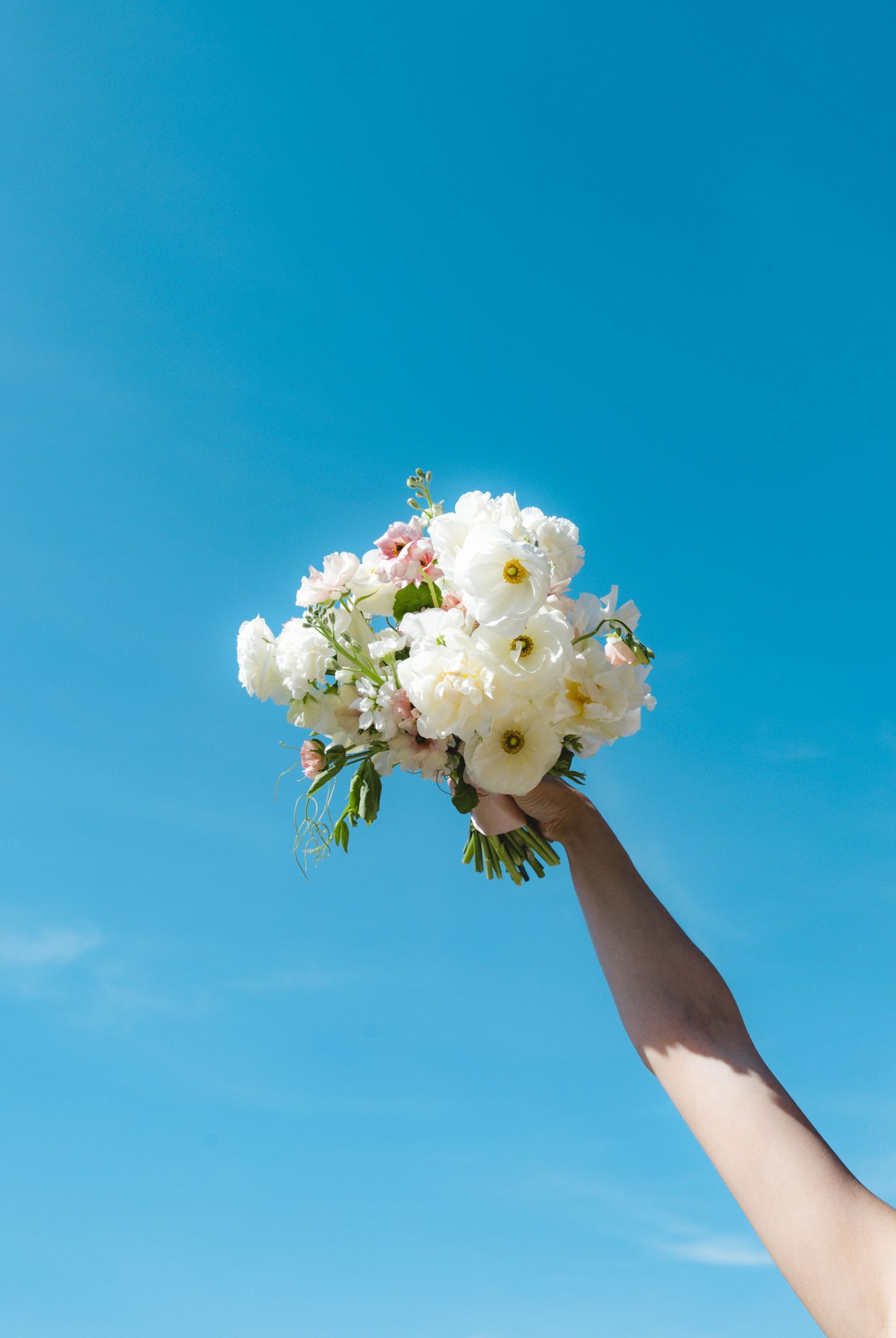 Hand holding a white wedding bouquet of peonies and roses raised against a bright blue sky.
