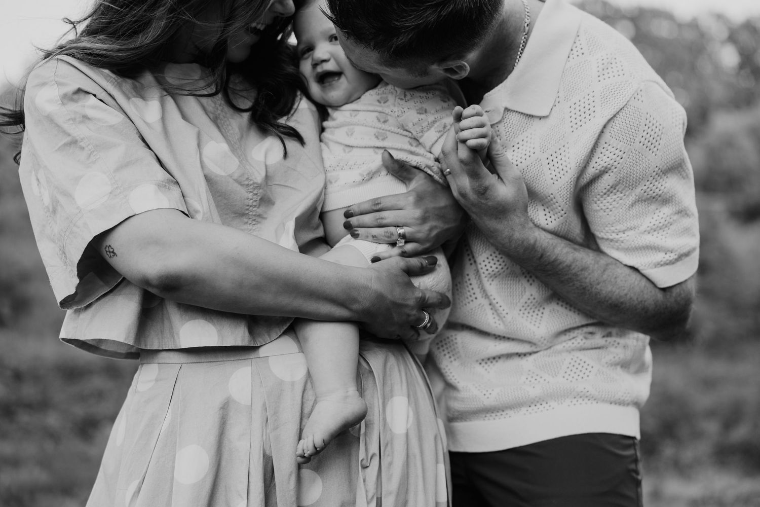 Black and white photo of parents lovingly holding their laughing toddler outdoors.