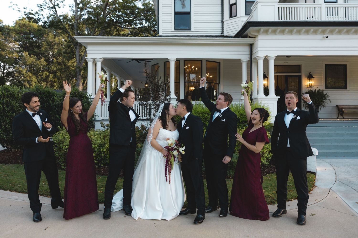Bride and groom kiss on a porch lawn while groomsmen in black tuxedos and bridesmaids in burgundy dresses cheer and raise their arms.
