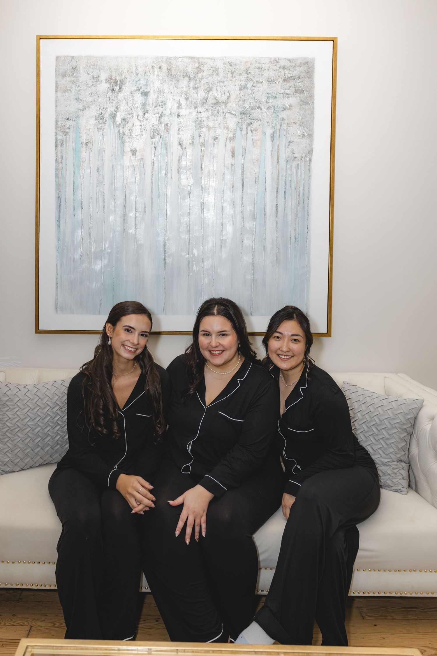 Three smiling women in matching black pajama sets sit together on a cream sofa beneath a large abstract blue and white painting.