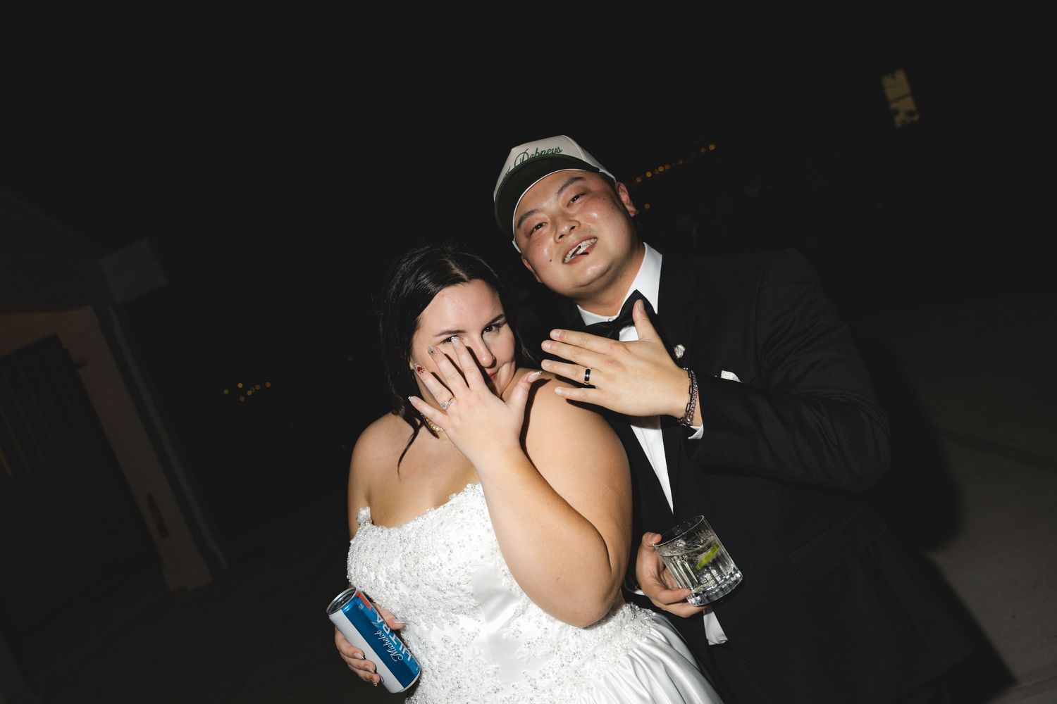 Bride in a strapless white wedding gown holds a drink and covers her face while a groom in a tuxedo and cap laughs beside her at night.