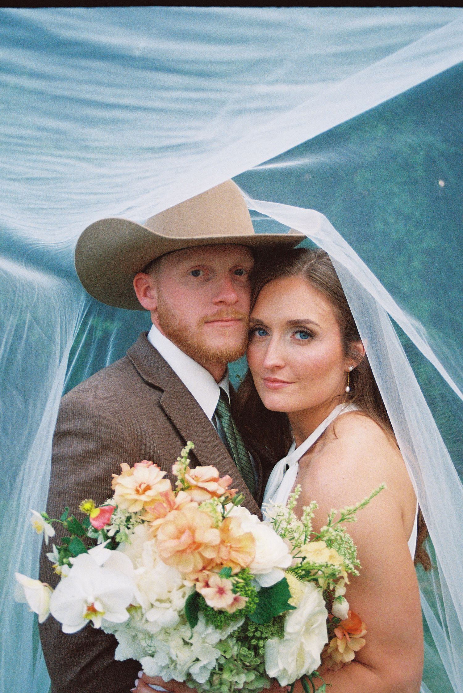 Bride and groom pose under a flowing blue veil, groom wearing a cowboy hat, bride holding a pastel floral bouquet.