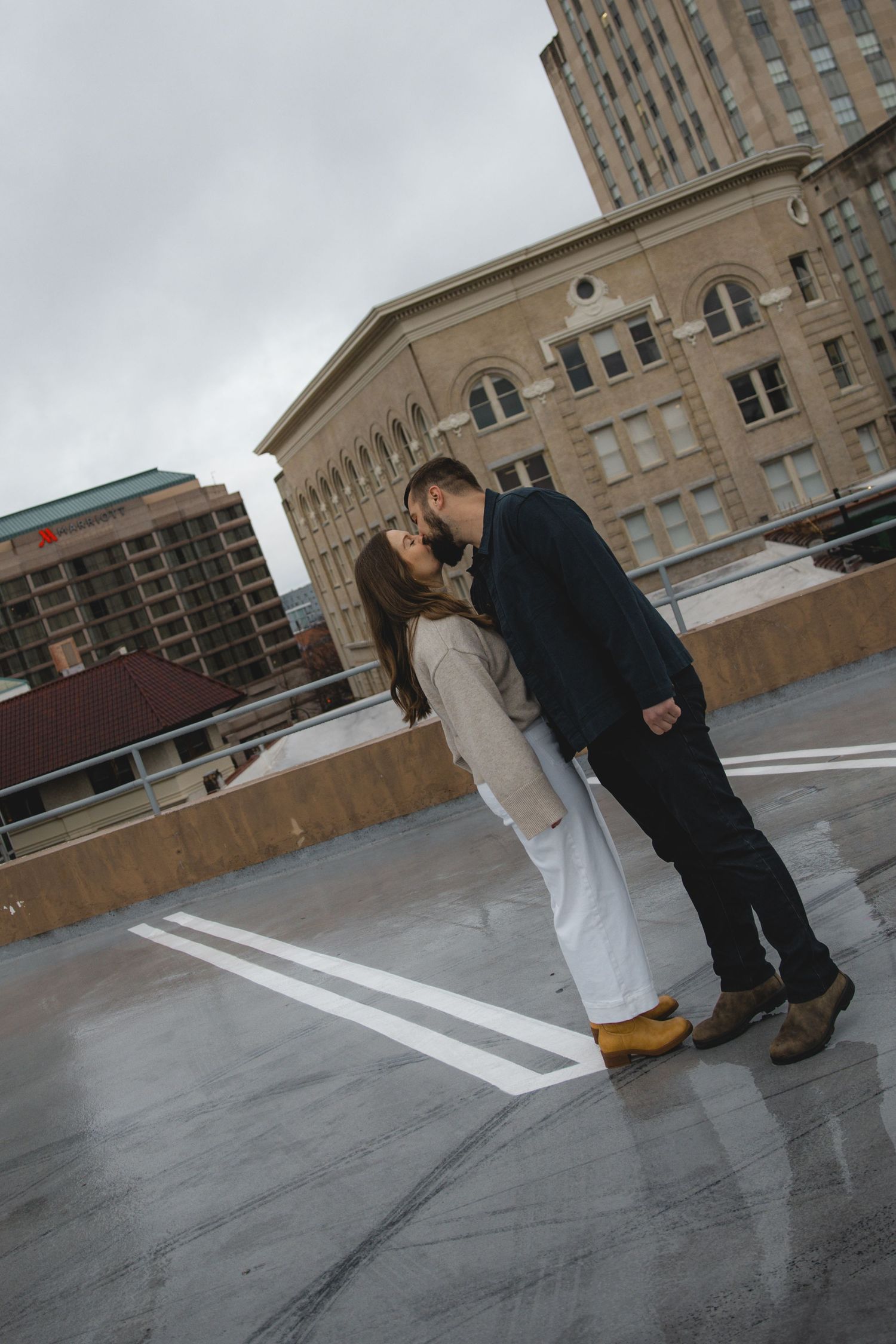 Couple sharing a romantic kiss on a rooftop parking garage with urban buildings in the background.