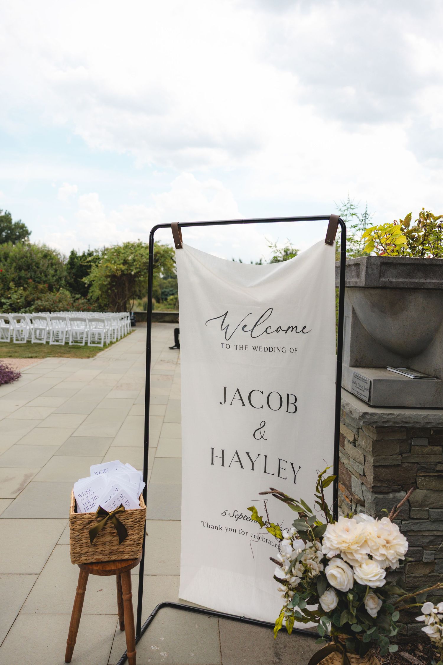 Elegant outdoor wedding welcome sign for Jacob and Hayley, surrounded by white floral arrangements and rows of chairs.