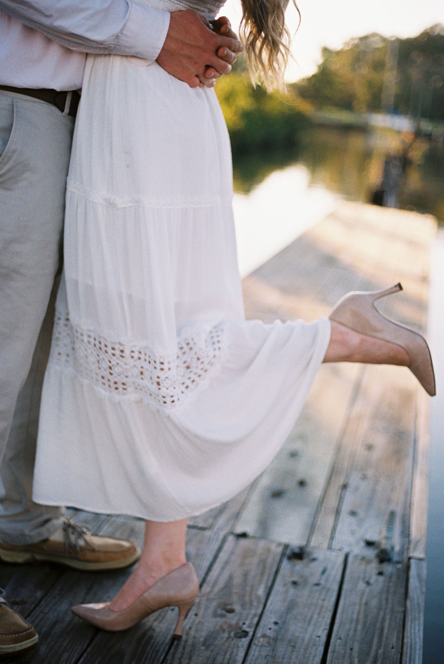 Couple embracing on a wooden dock, woman in white lace maxi skirt and nude heels, man in gray pants and boat shoes.
