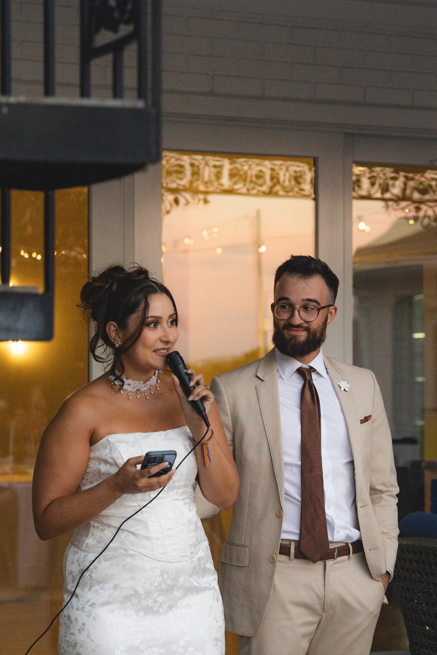 Bride in white strapless gown speaks into microphone beside groom in tan suit with brown tie at wedding reception.