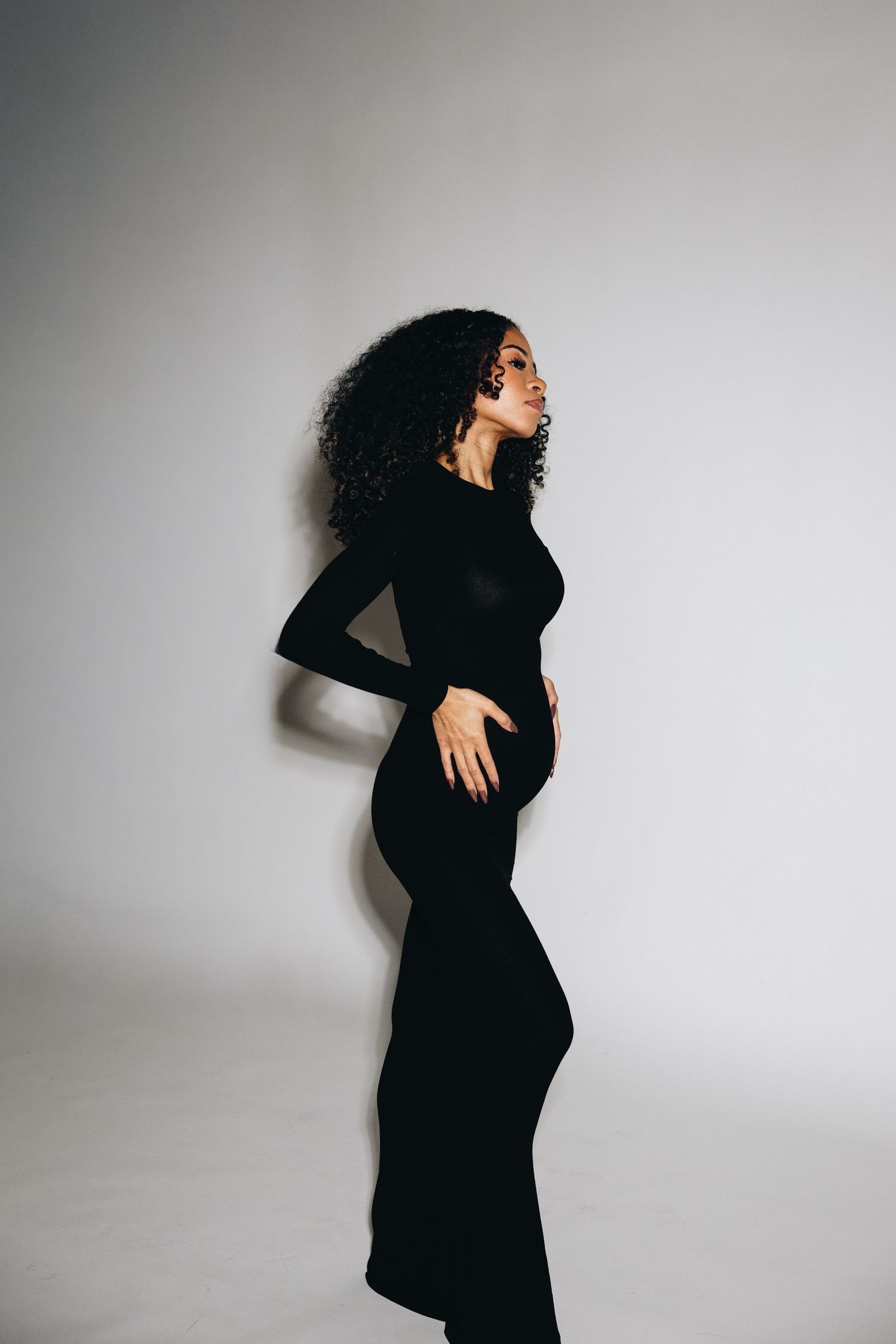 A woman in a black long-sleeve fitted maxi dress poses sideways against a white background.