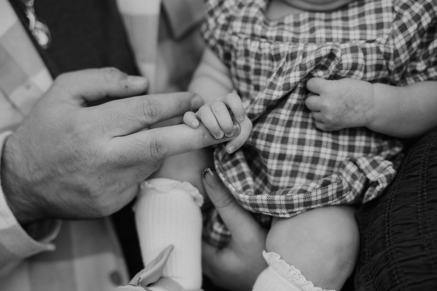 Black and white photo of an adult gently holding a newborn baby's tiny hand, infant wearing plaid outfit.