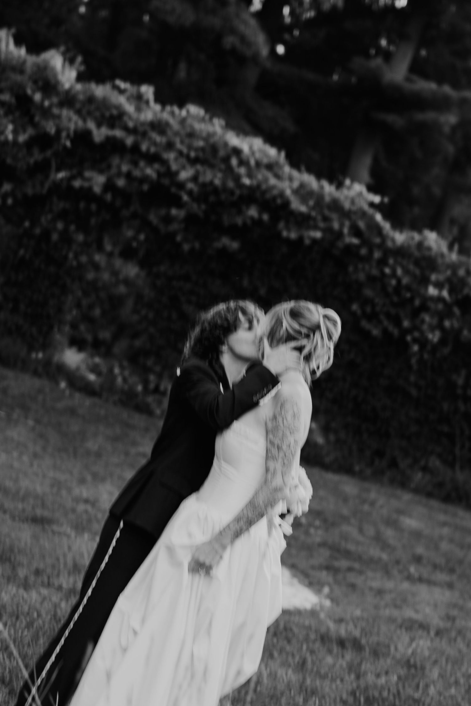 Black and white photo of a bride and groom sharing a romantic kiss outdoors on a lush green lawn.