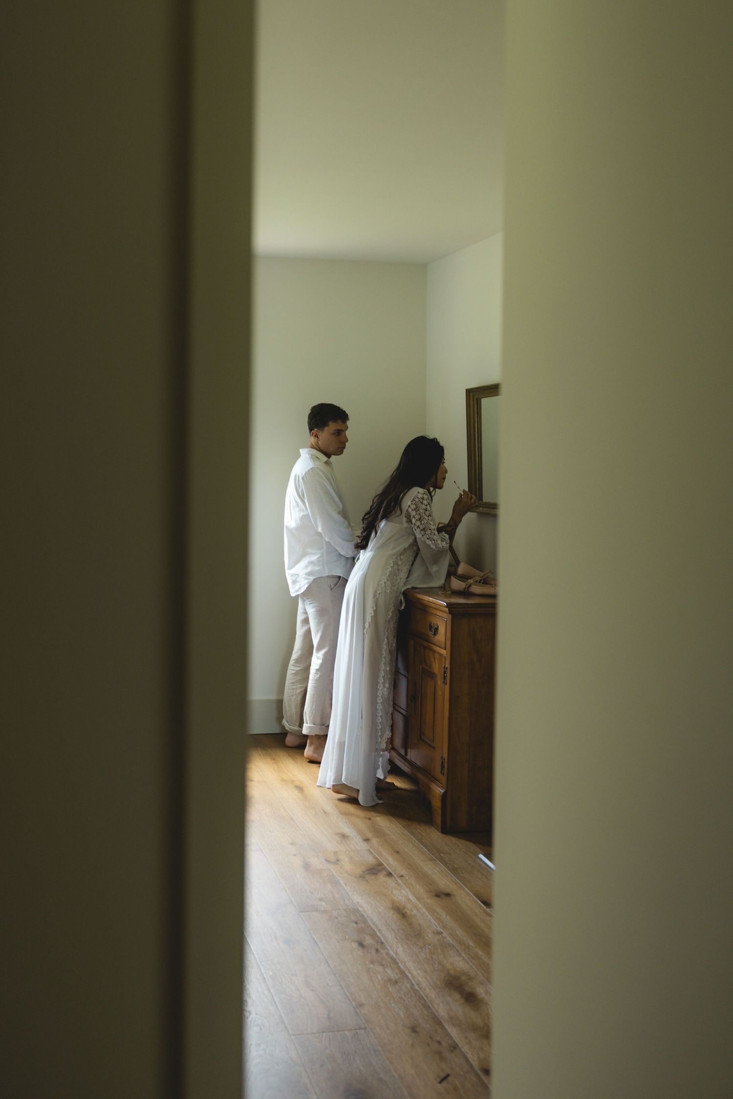 A couple dressed in white stands together in a sunlit room with wooden floors, viewed through a doorway.