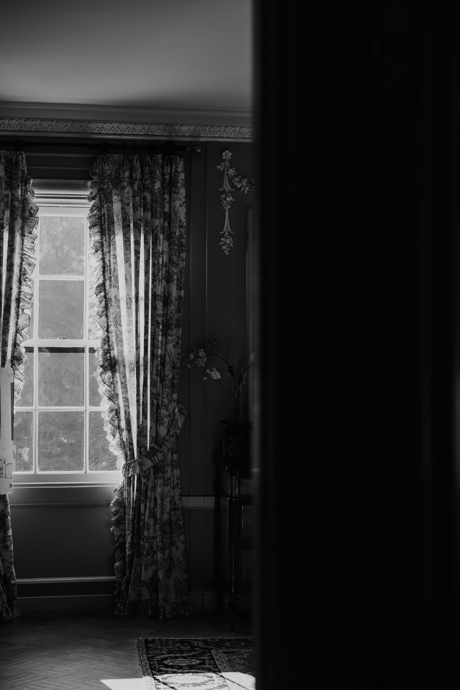Black and white photo of an elegant room with ornate curtains framing a tall window, casting soft light on a patterned rug.