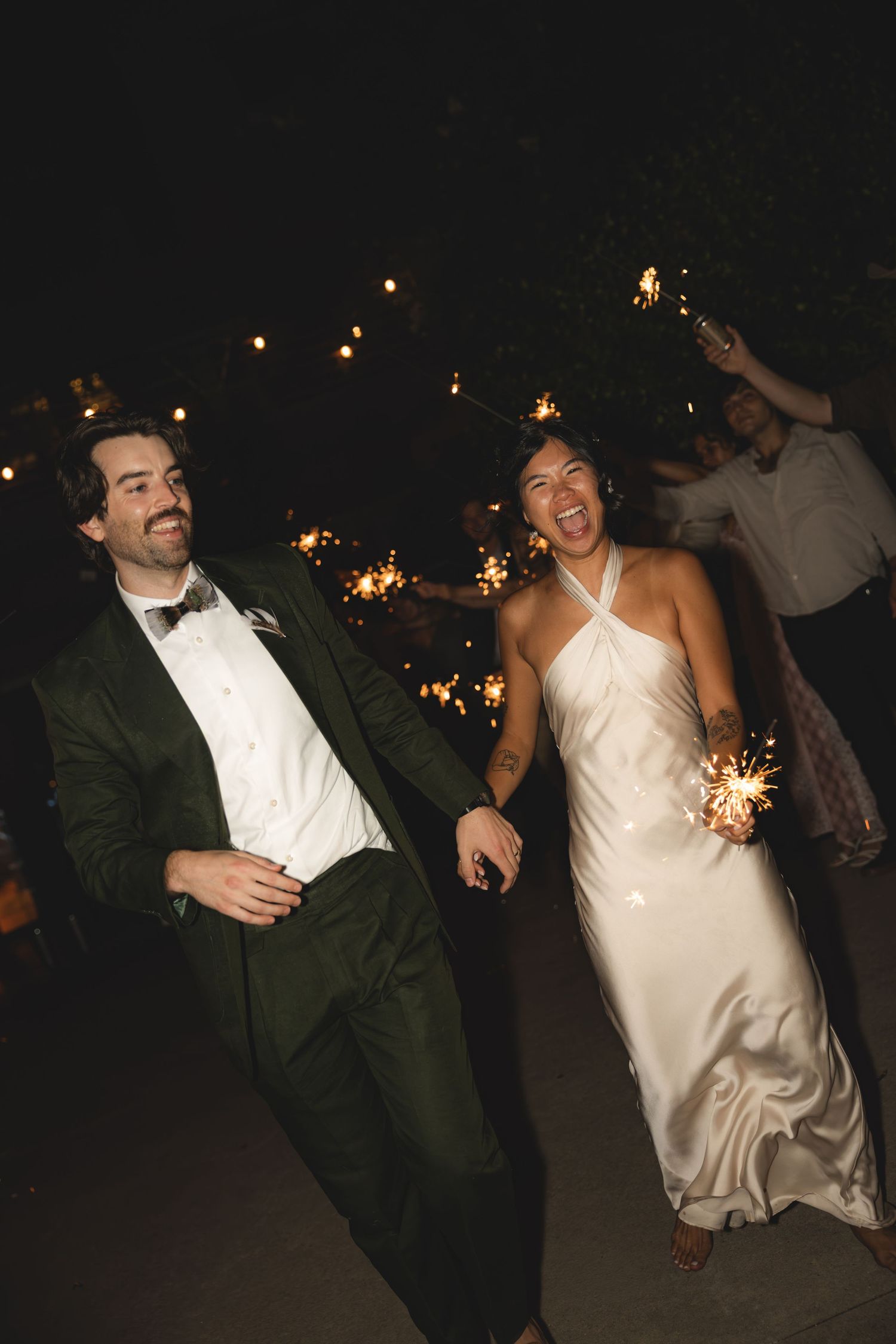 Joyful newlyweds walk through a sparkler send-off at night, bride in white satin gown, groom in dark green tuxedo.