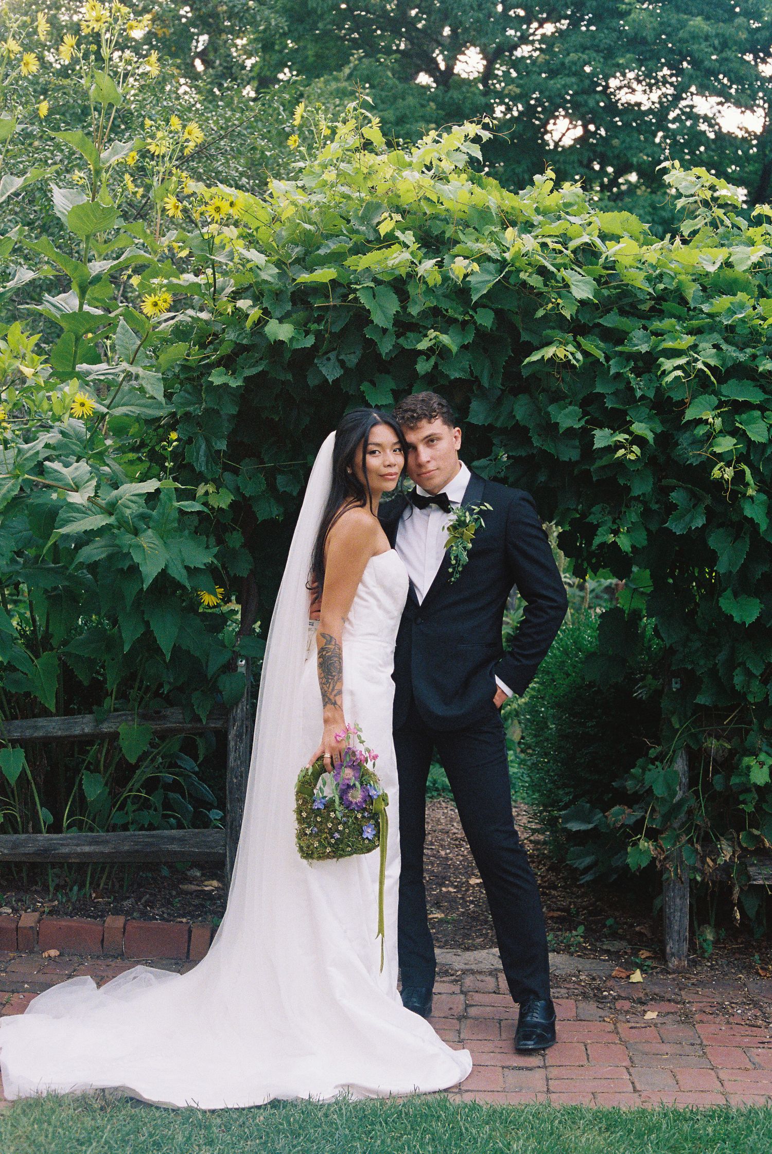 Bride in white gown with veil and groom in black tuxedo pose together in lush garden on their wedding day.