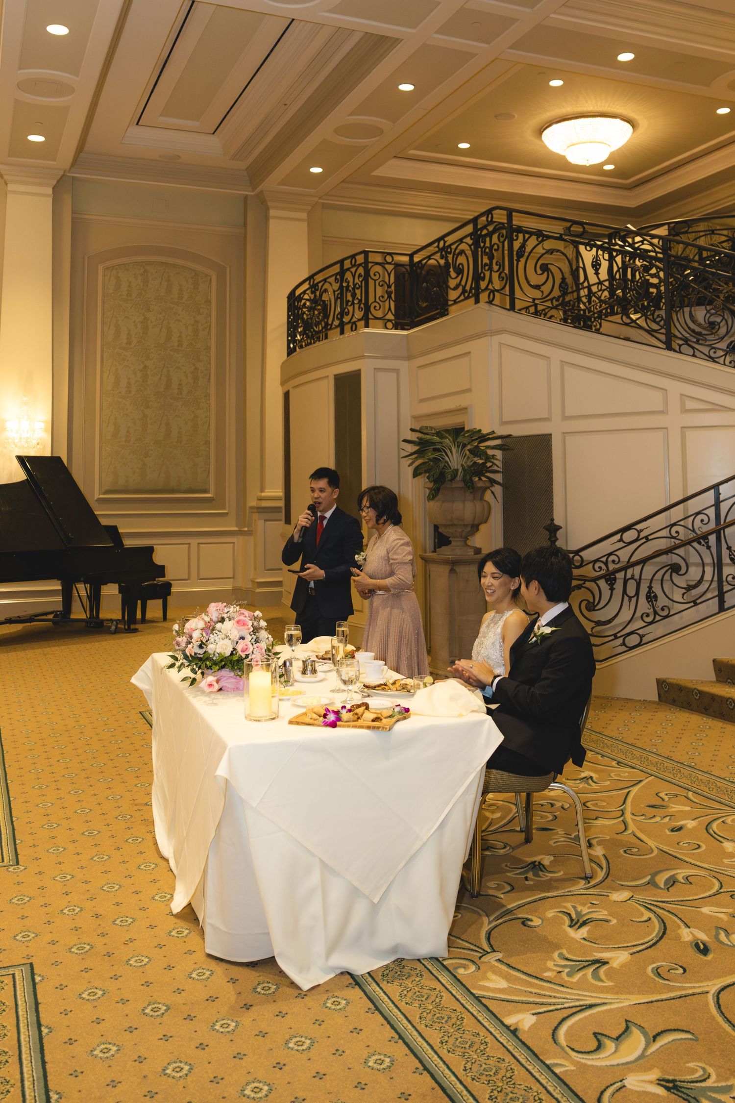 Couple seated at sweetheart table during wedding reception toast in grand ballroom.